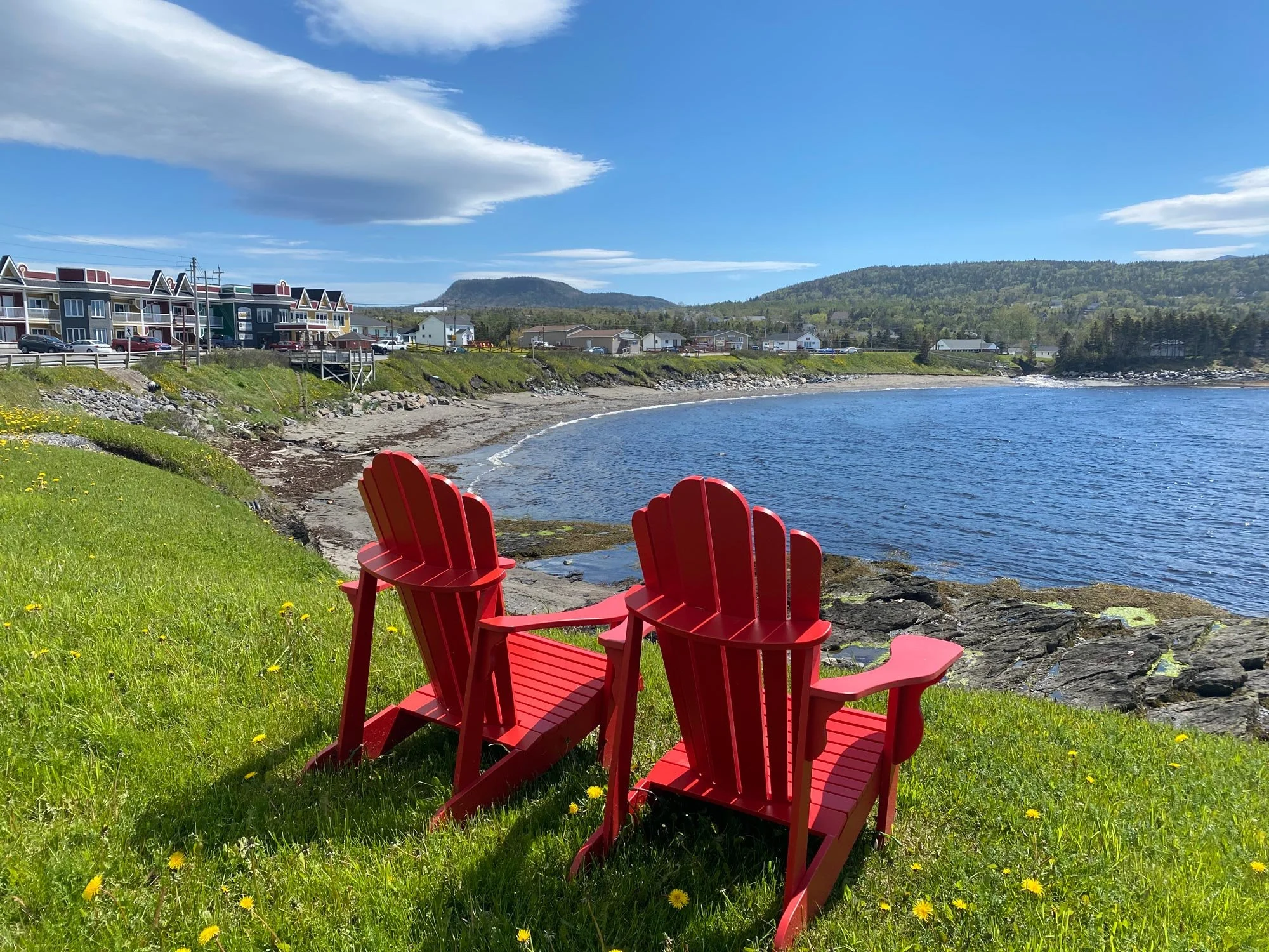 Two red Adirondack chairs on green grass by a rocky shoreline, overlooking a calm body of water under a blue sky with scattered clouds, with colorful houses and hills in the background.