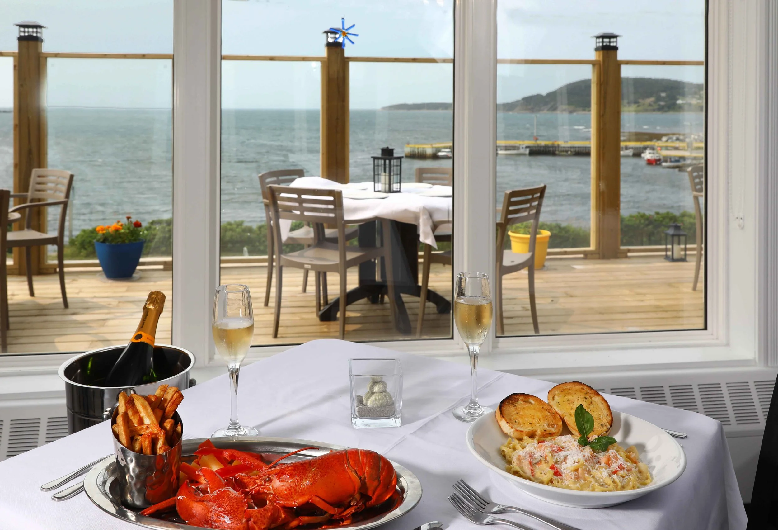 A table set with lobster, French fries, pasta with seafood, champagne, and garlic bread in front of a window overlooking a dock and body of water.