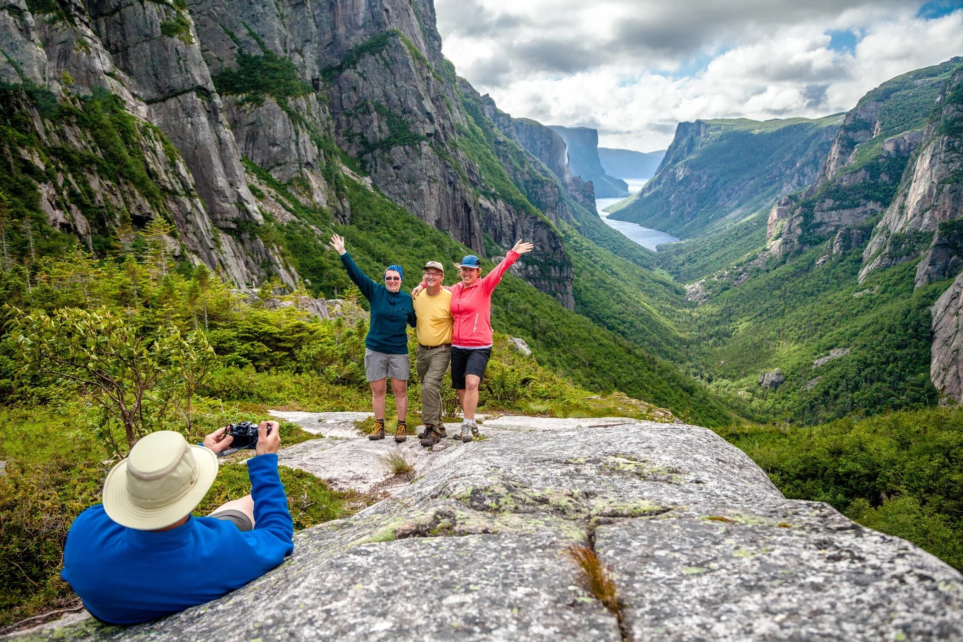 Three people standing on a rock in a lush, green valley with tall cliffs on either side, taking a photo of a person sitting with a camera on the ground in front of them, all enjoying the scenic view of the valley and river.