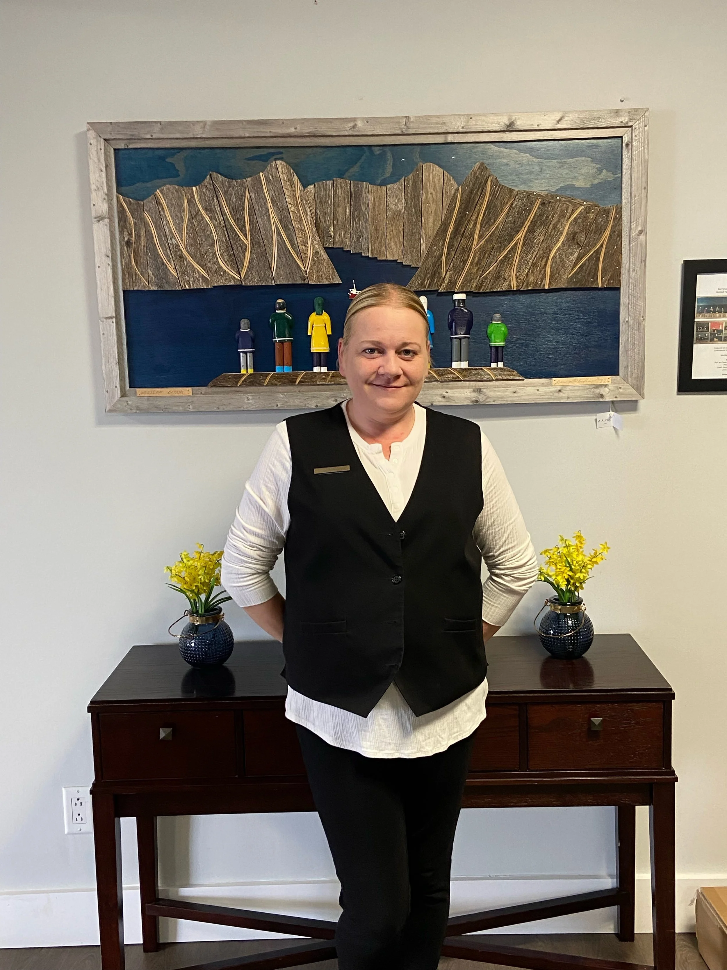 A woman standing in front of a dark wooden table with yellow flowers in vases, in a room with a framed art piece on the wall depicting colorful wooden figures by a lake and mountains.