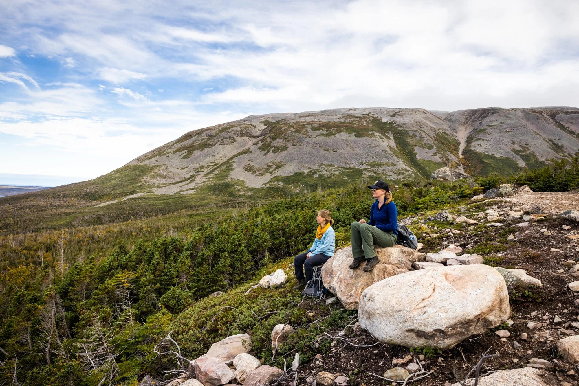 Two women sitting on large rocks on a mountain trail, overlooking a forested valley with a mountain in the background during daytime.