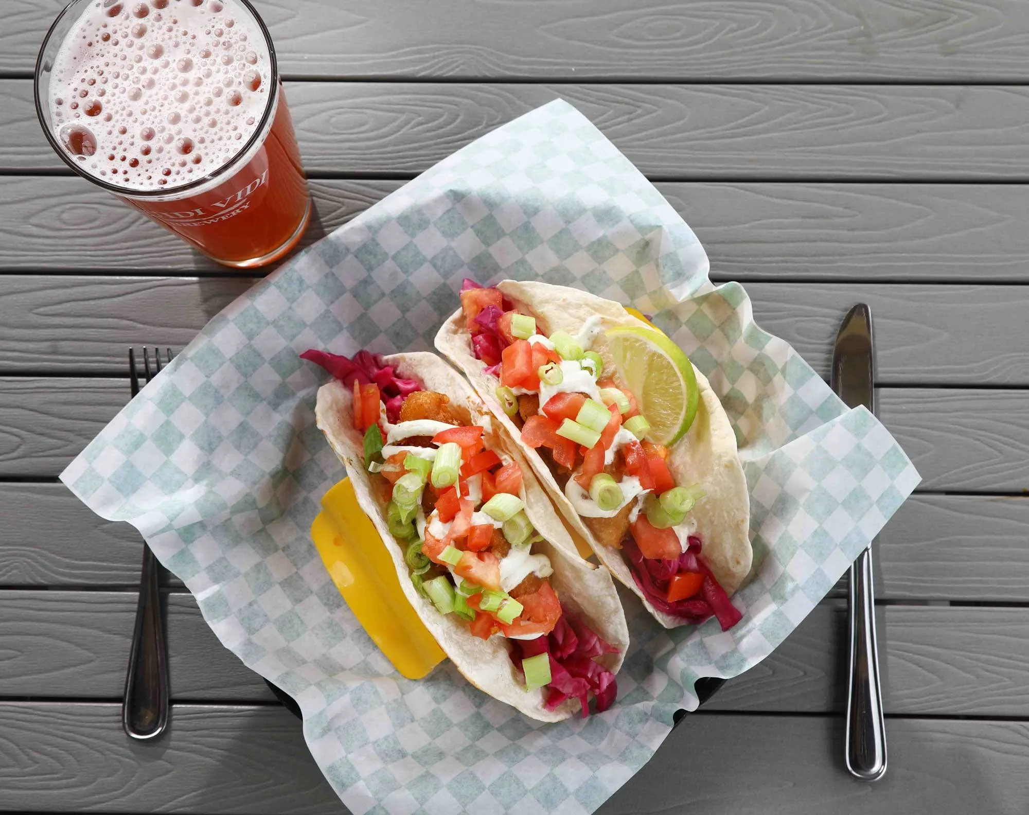 Two fish tacos topped with diced tomatoes, green onions, and sauce, served with lemon wedges on a checkered paper-lined basket, accompanied by a slice of yellow cheese, a slice of lemon, and a glass of reddish beverage on a gray wooden table with a f