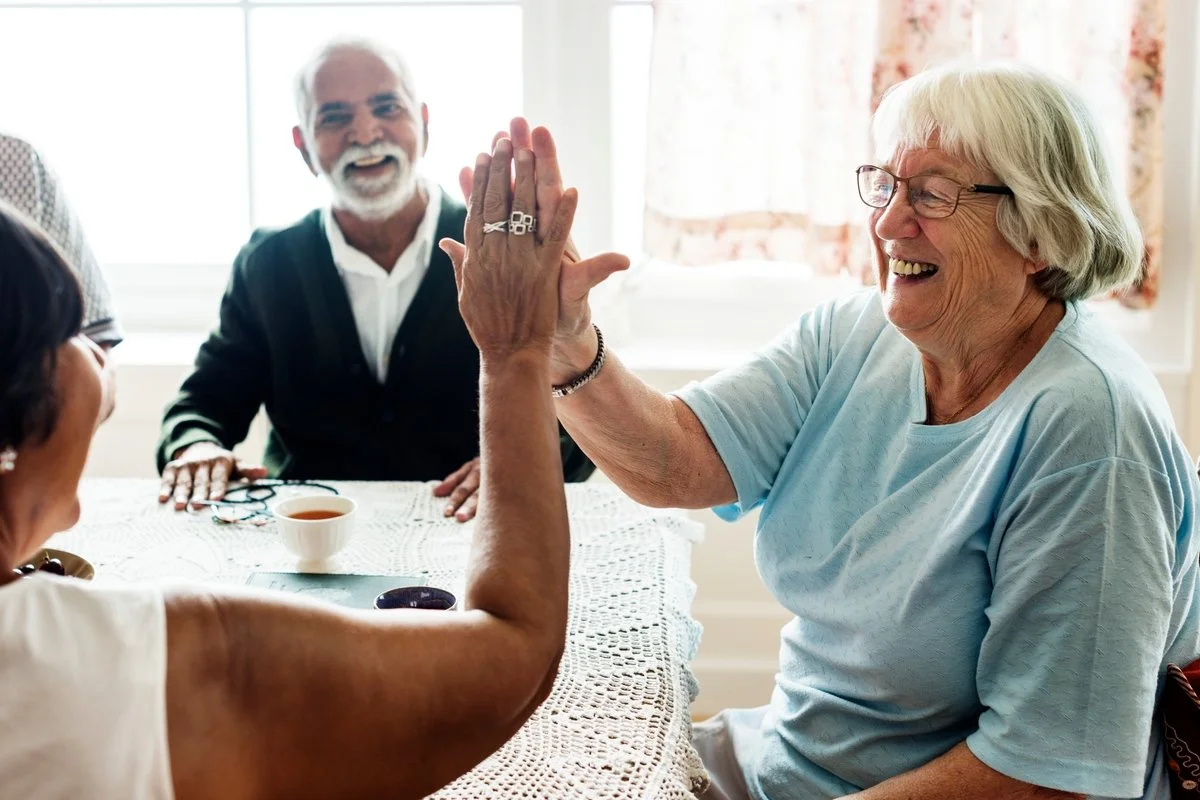 Senior women playing high five with a man sitting at a table, smiling, in a bright room.