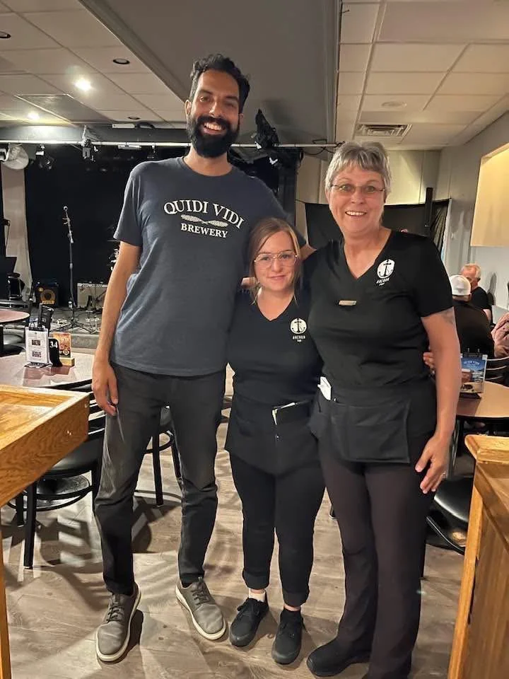 Three people standing together in a restaurant or bar, smiling at the camera. Two women in black uniforms and a tall man in a gray t-shirt with 'Quidi Vidi Brewery' written on it.