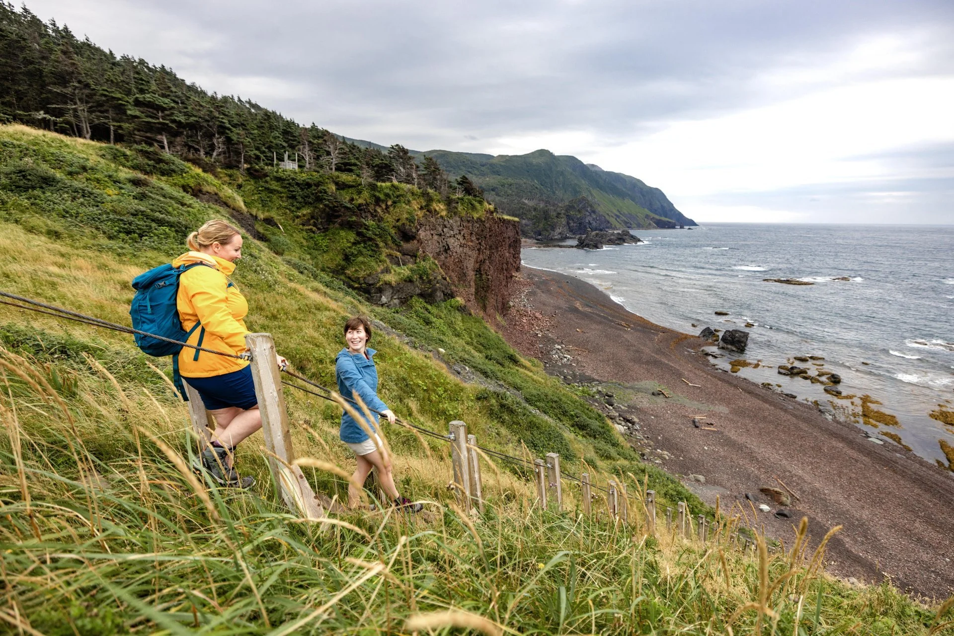Two women hiking along a coastal trail with cliffs, grassy terrain, and the ocean in the background.