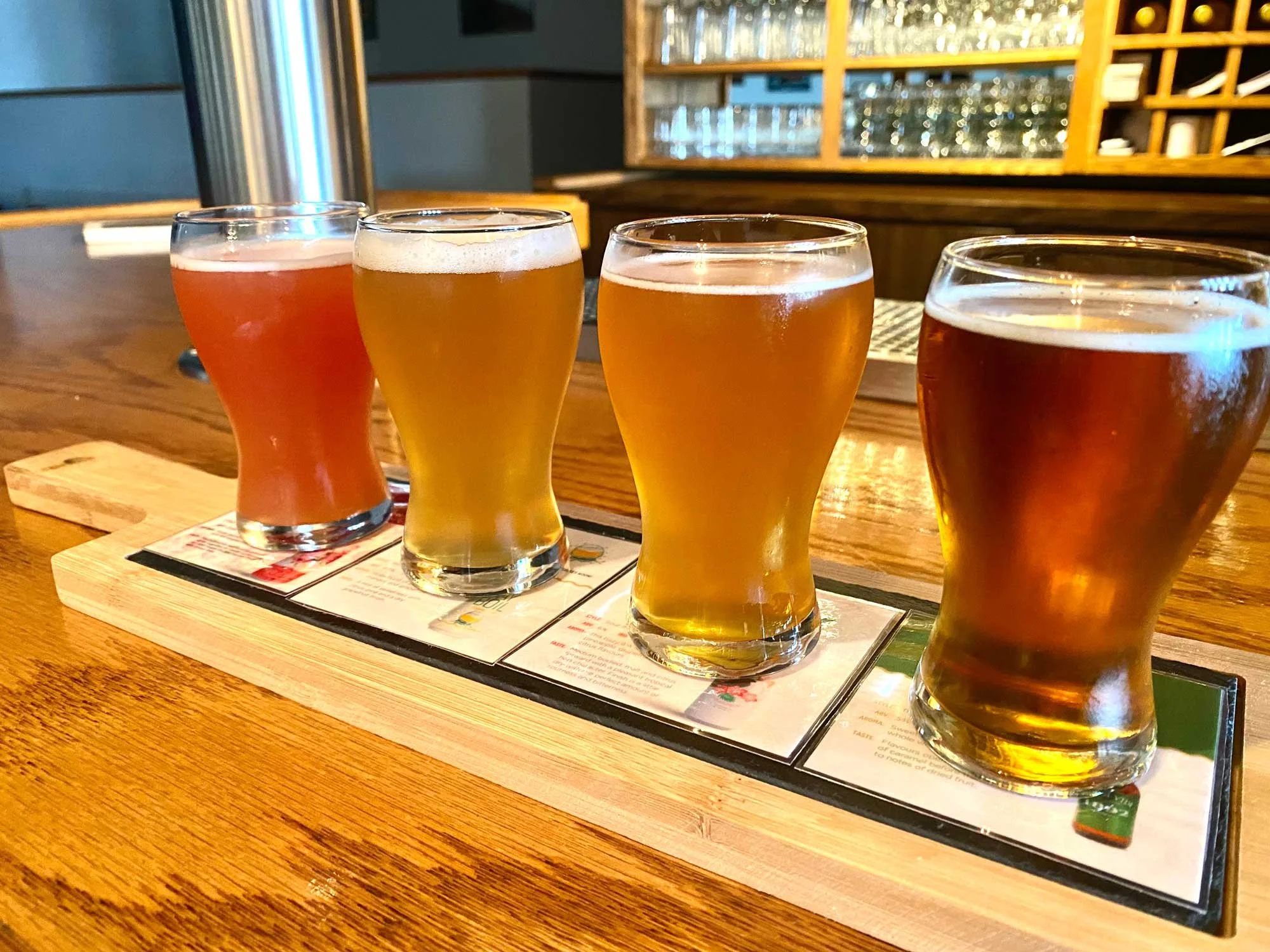 A flight of four different beers served in small, curvy glasses on a wooden flight paddle, placed on a menu on a wooden table in a bar or brewery setting.