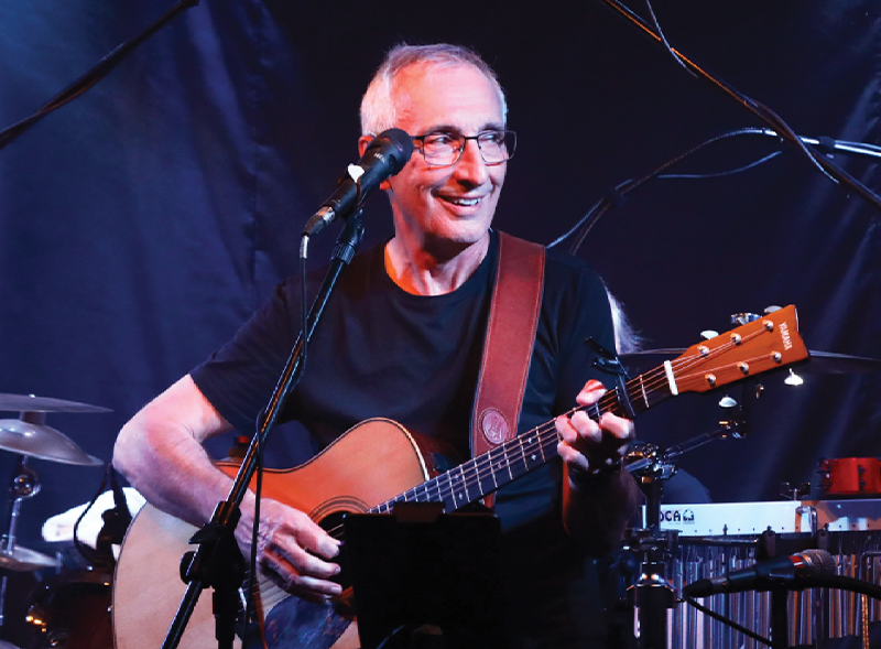 A man playing an acoustic guitar on stage, smiling, with a microphone in front of him, and musical equipment in the background.