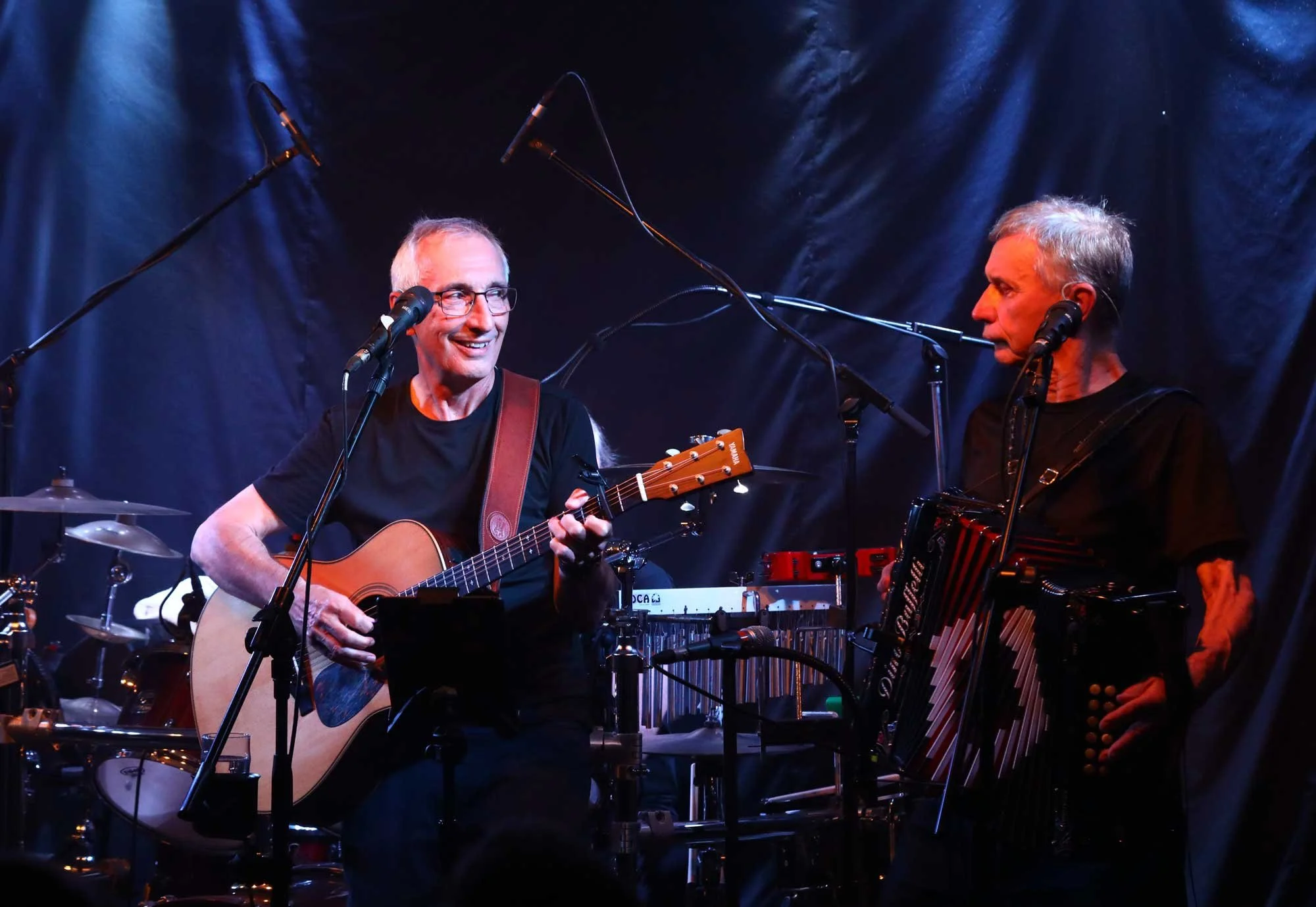 Two male musicians performing on stage, one playing an acoustic guitar and the other with an accordion, with microphones and sound equipment around them.
