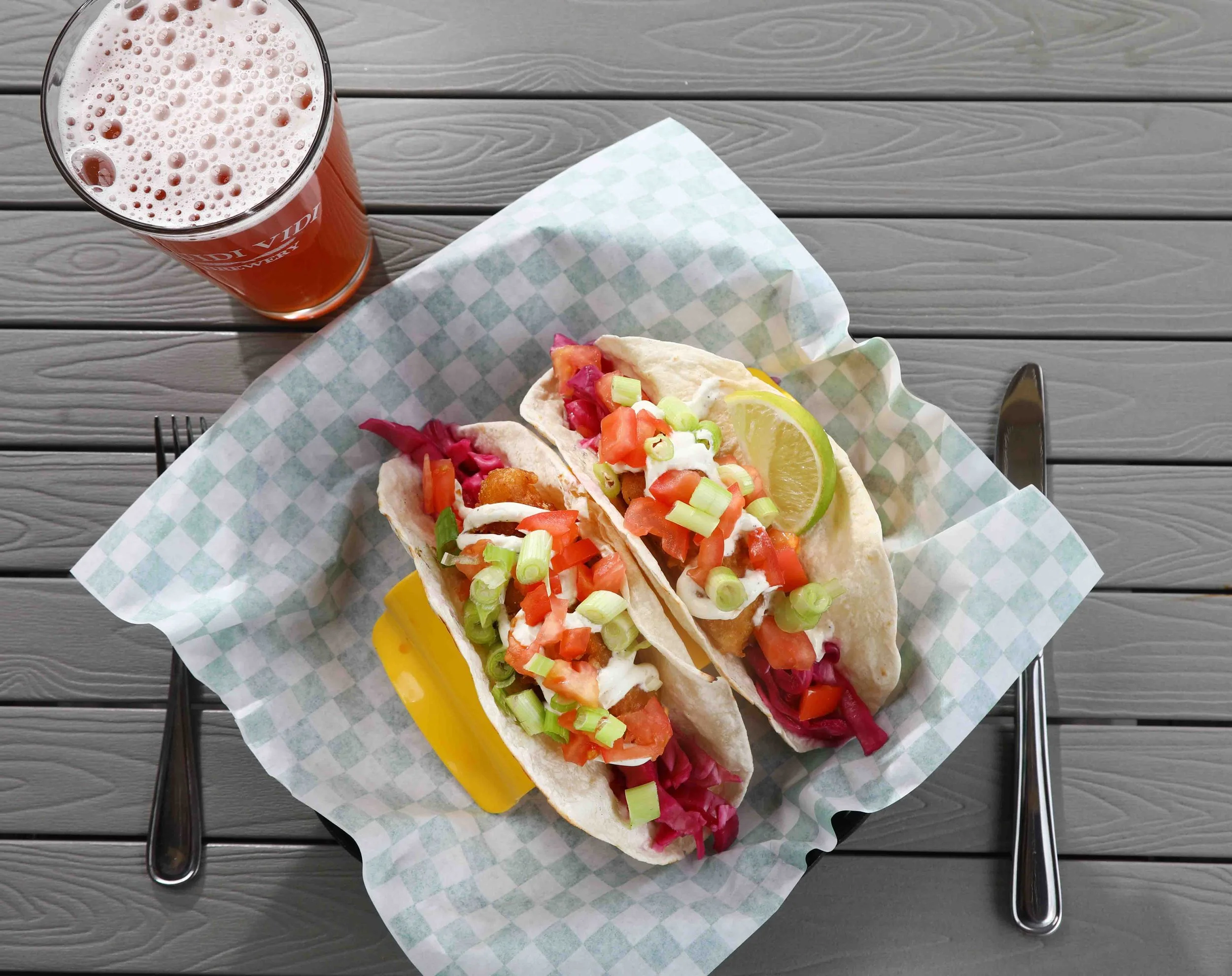 Two fish tacos topped with chopped tomatoes, green onions, and sour cream, served with a lemon wedge and a side of yellow mustard, accompanied by a glass of amber-colored beer on a wooden table.