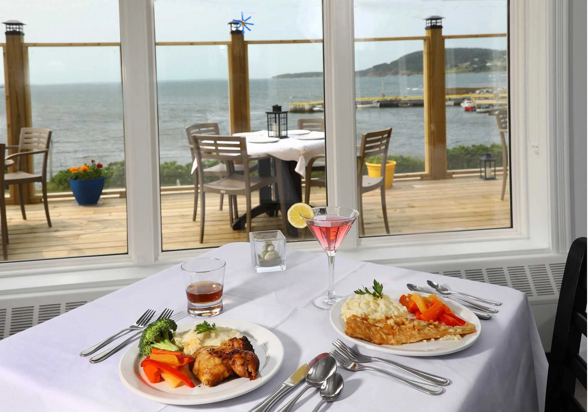 Indoor dining table set with plates of food, including mashed potatoes, vegetables, and grilled chicken, with drinks, in front of large window showing a wooden deck outside overlooking water and boats, with a dining area and landscape in the backgrou