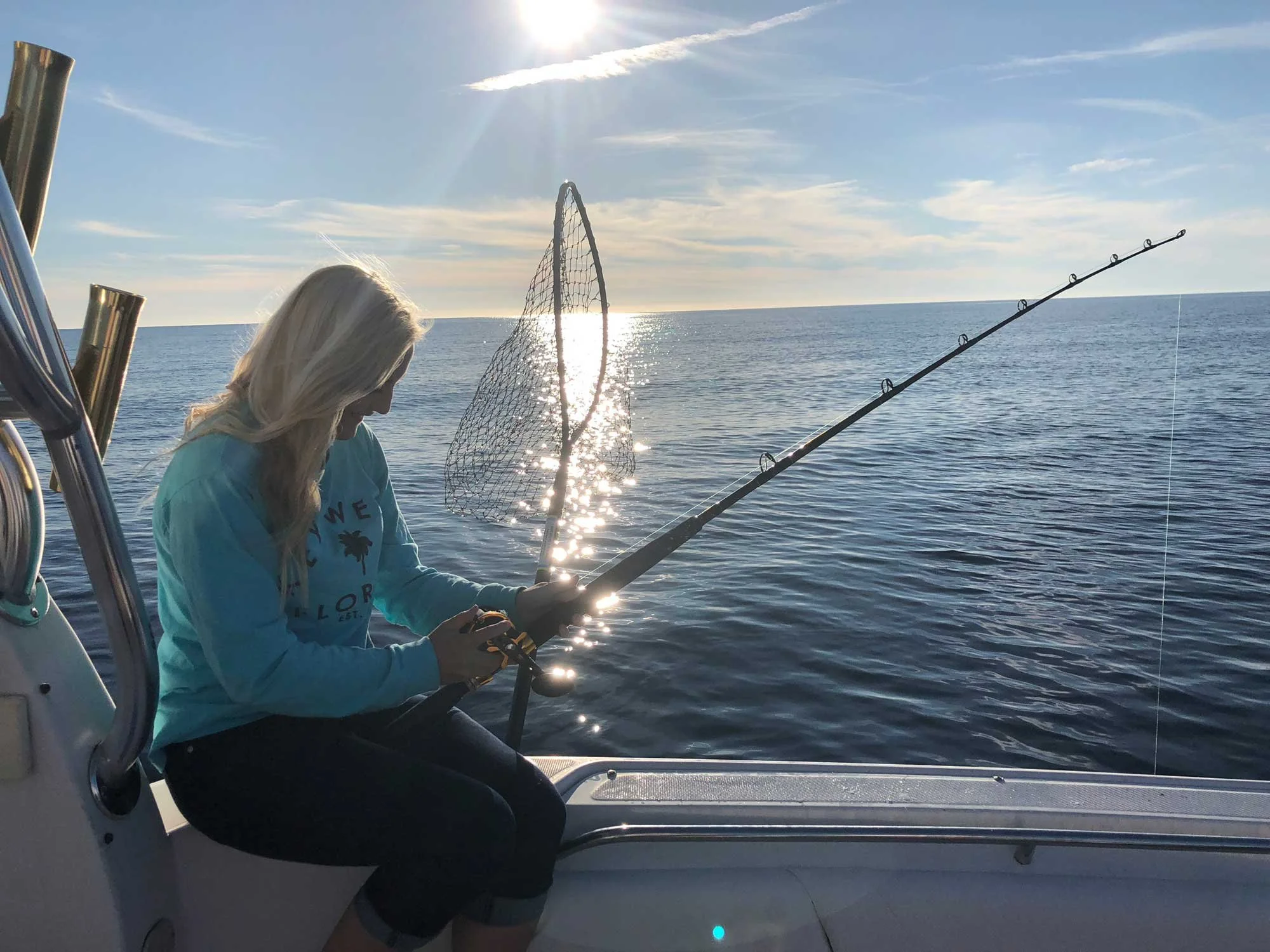 Woman sitting on a boat holding a fishing rod with ocean and sunset behind her.