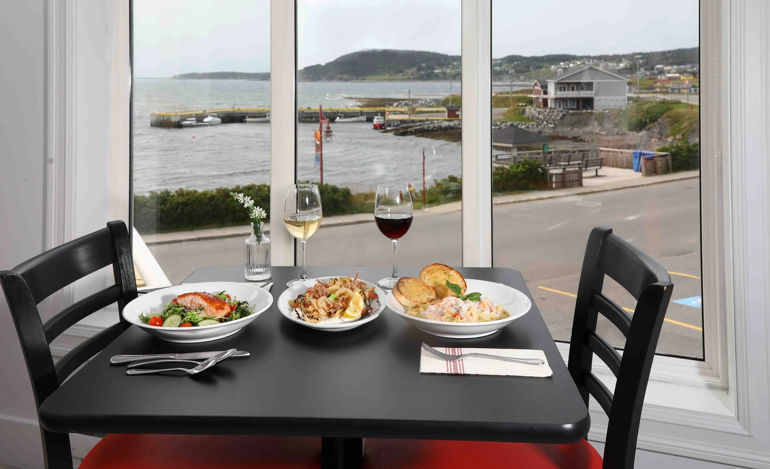 A dining table set with three plates of food, two glasses of wine, and a small vase with white flowers. The background shows a large window overlooking a harbor with water, boats, and hills.
