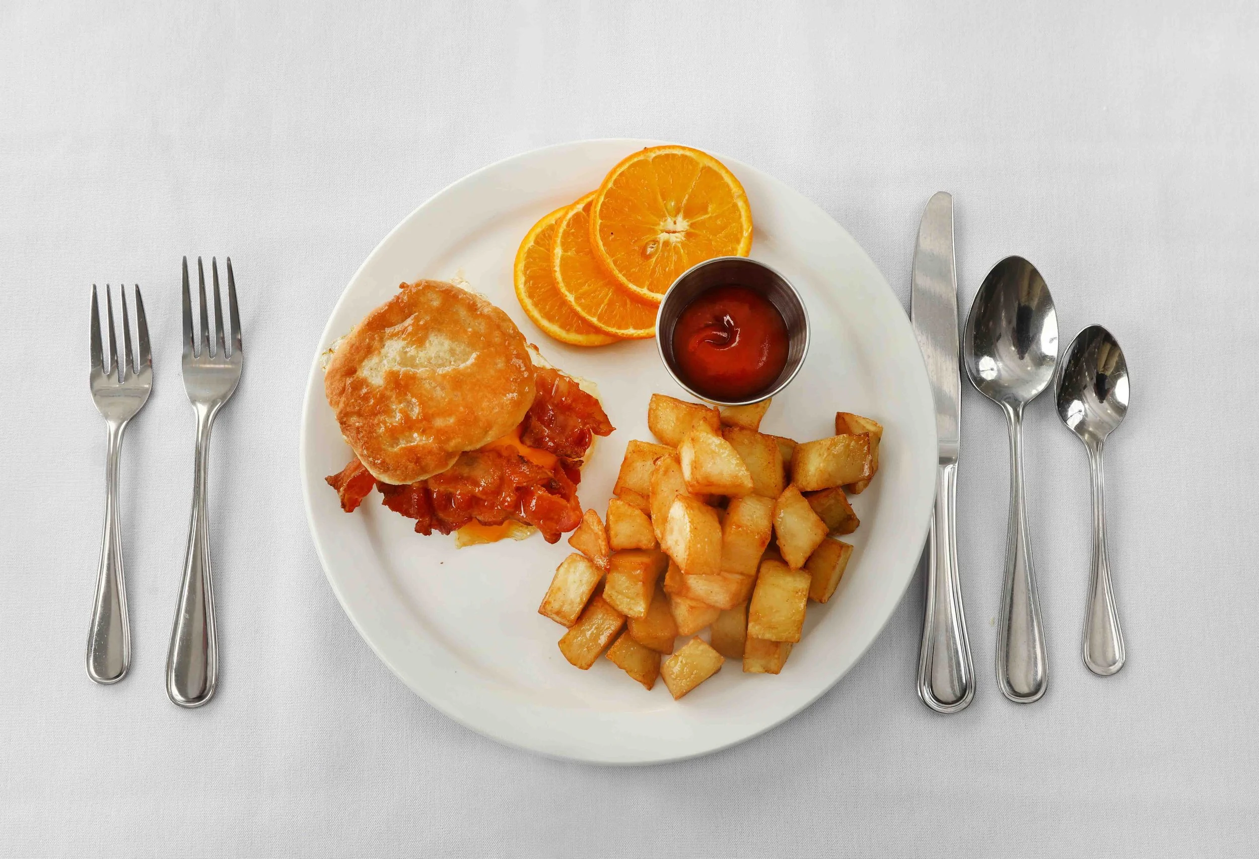 Plate with chicken and waffles, potato cubes, sliced orange, and a small cup of ketchup on a white tablecloth, with forks, knives, and spoons arranged around.