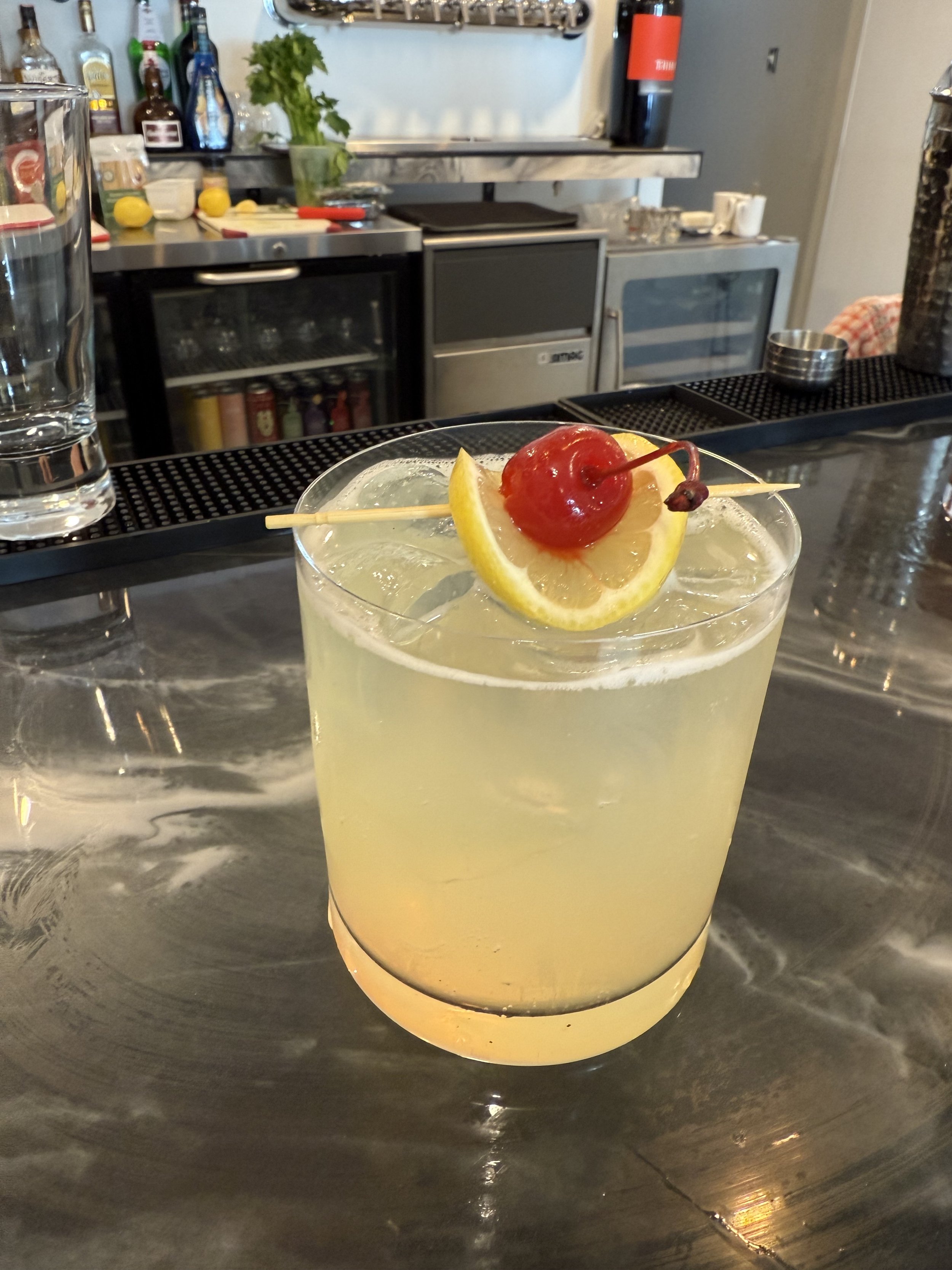 A cocktail garnished with a lemon slice and a maraschino cherry with a toothpick, on a bar counter.