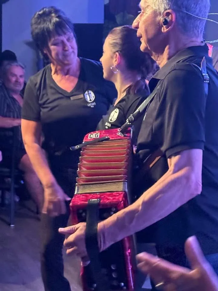 Three people, two women and a man, are standing close together at what appears to be a social gathering or party. The woman on the left has short dark hair and is smiling, wearing a black T-shirt with a circular logo. The woman in the middle has her 