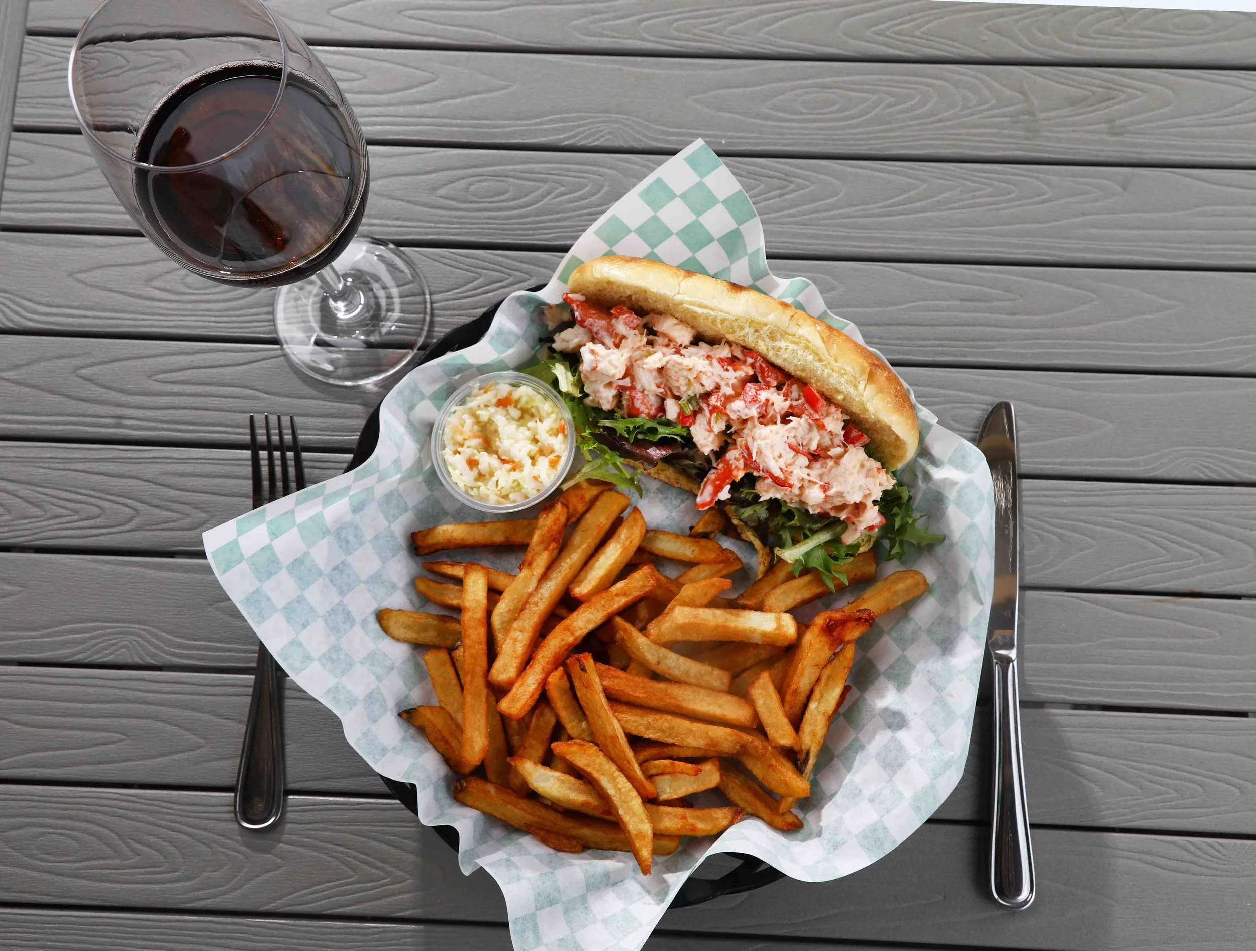 Lobster roll sandwich with lettuce, served with French fries, cole slaw, and a glass of red wine on a gray wooden table.
