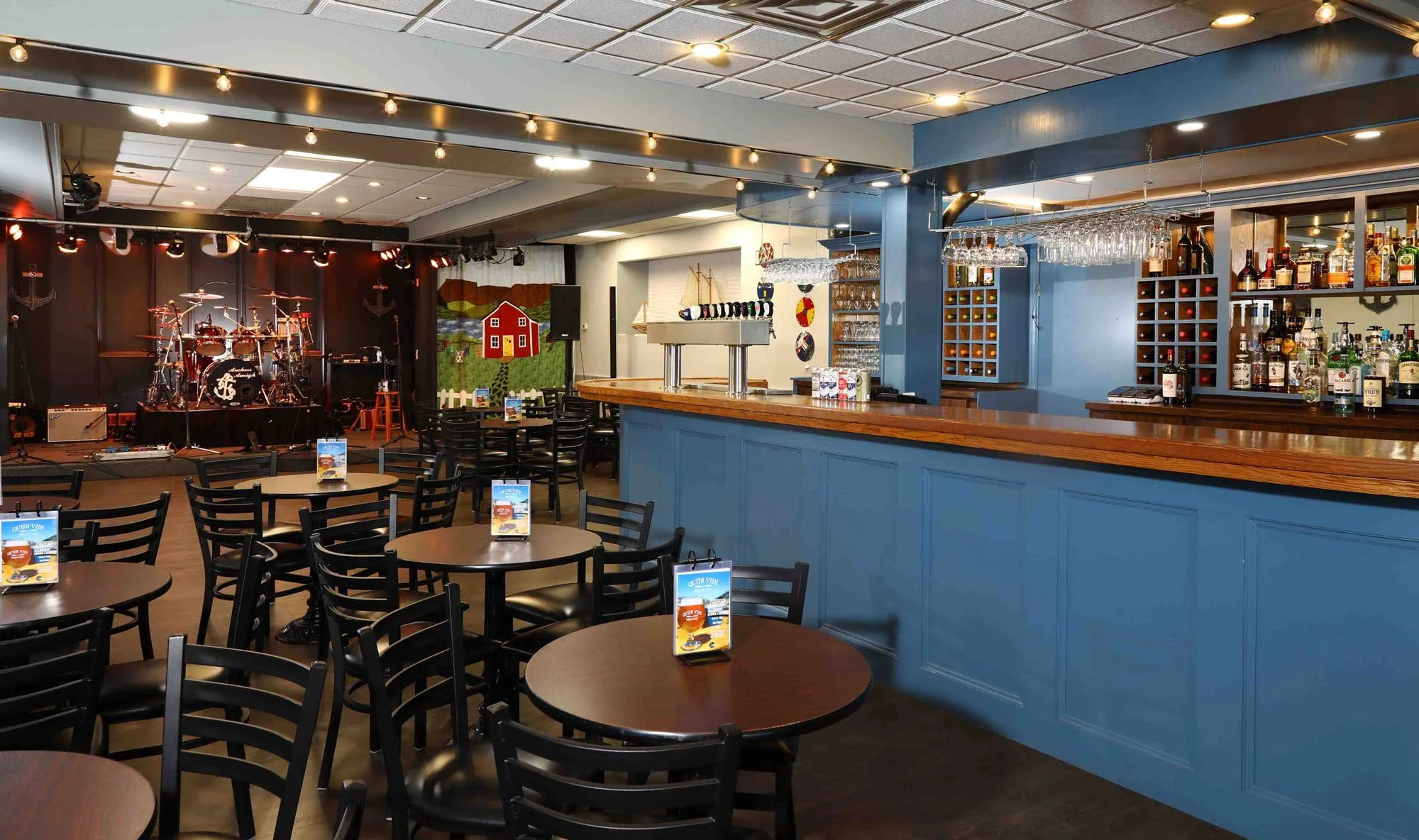 An indoor music venue with round tables and chairs, a bar area with blue cabinetry, and a small stage with musical instruments.