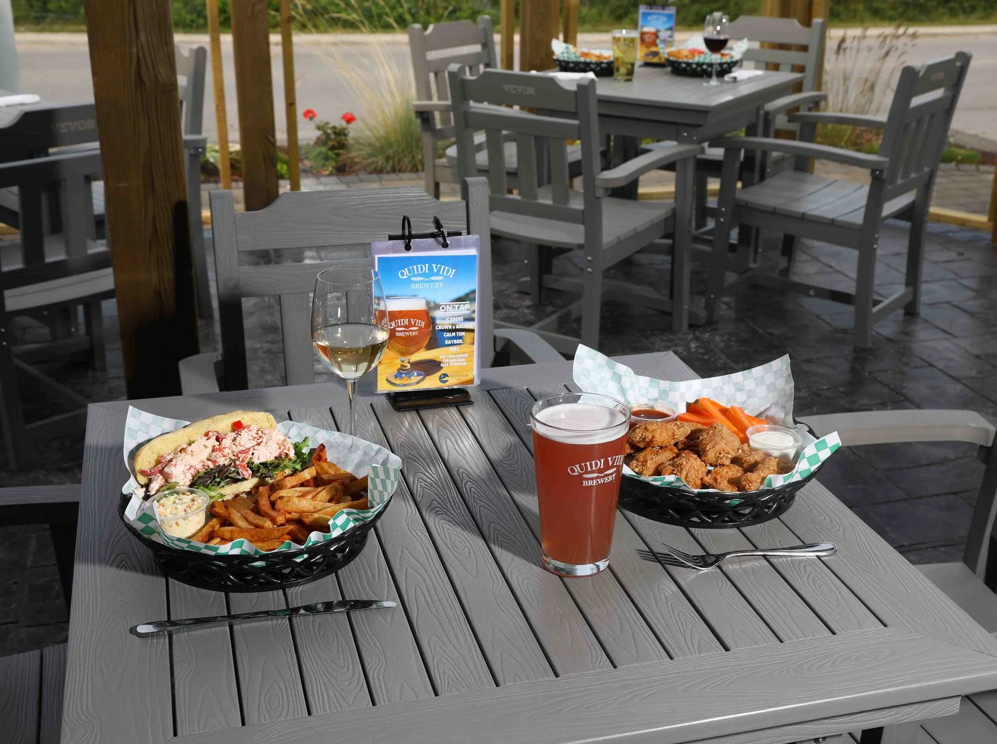Outdoor patio dining table with fried chicken, french fries, lobster roll, cold drinks, and a beer on a menu stand, with additional empty chairs and tables in the background.