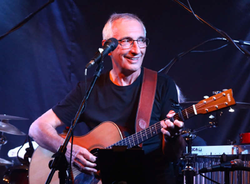 A man with short gray hair and glasses playing an acoustic guitar on stage, smiling, with a microphone in front of him, and musical equipment in the background.