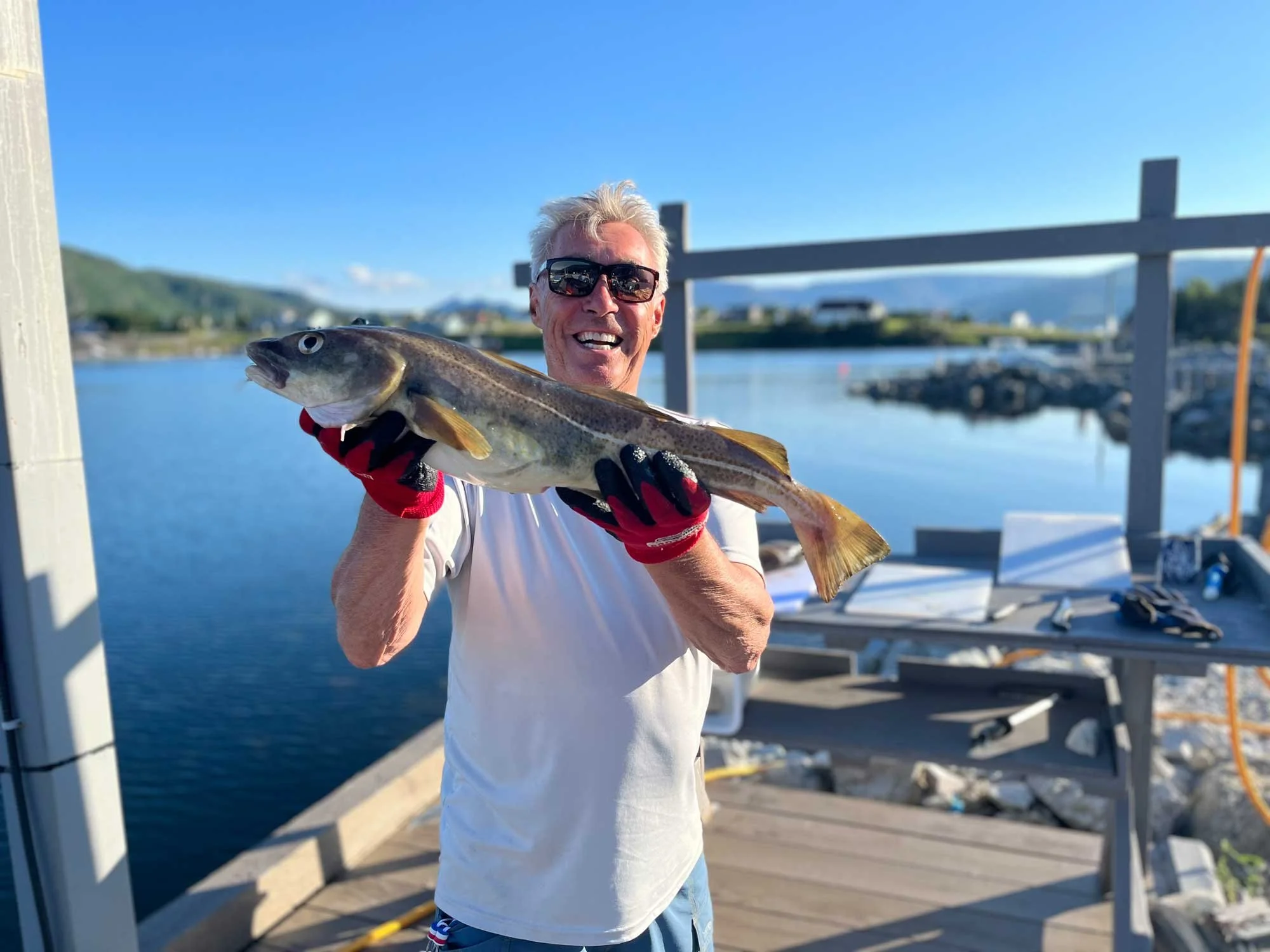 A man with white hair wearing sunglasses, a white shirt, and red gloves is smiling while holding a large fish he caught at a dock by the water.
