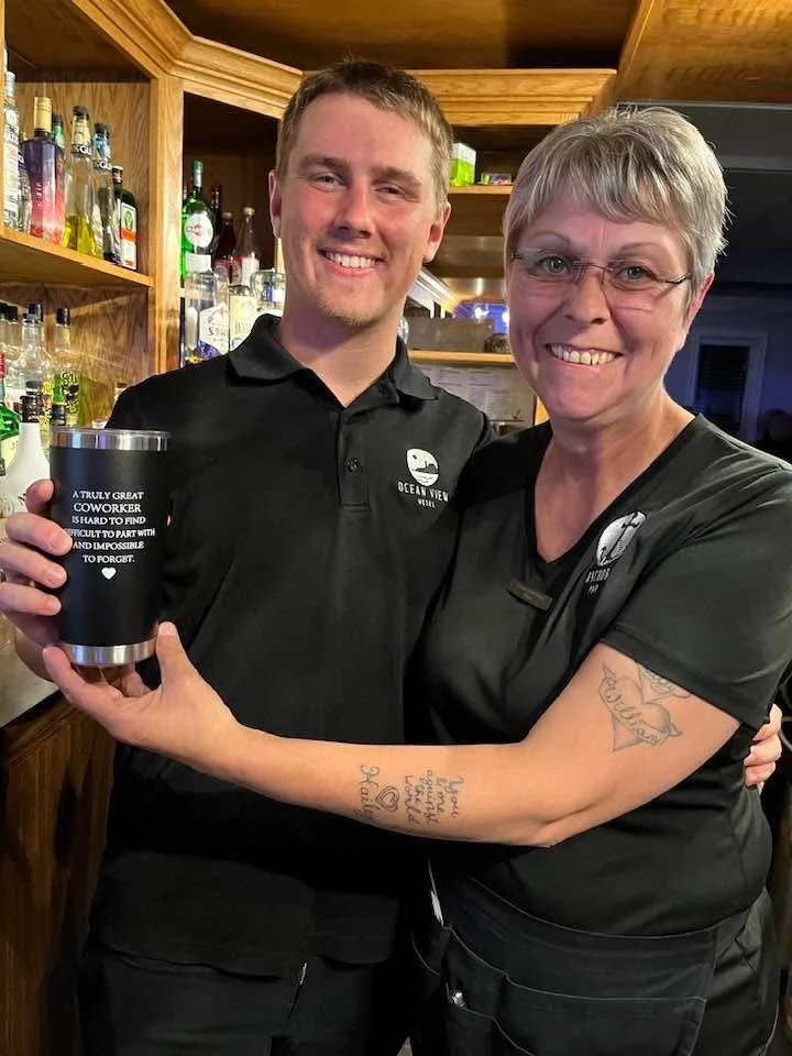 A young man and a woman, both smiling, embrace inside a bar or restaurant. The young man holds a black tumbler with text on it. Both are wearing black shirts with a logo for Ocean View. The woman has tattoos on her arm.