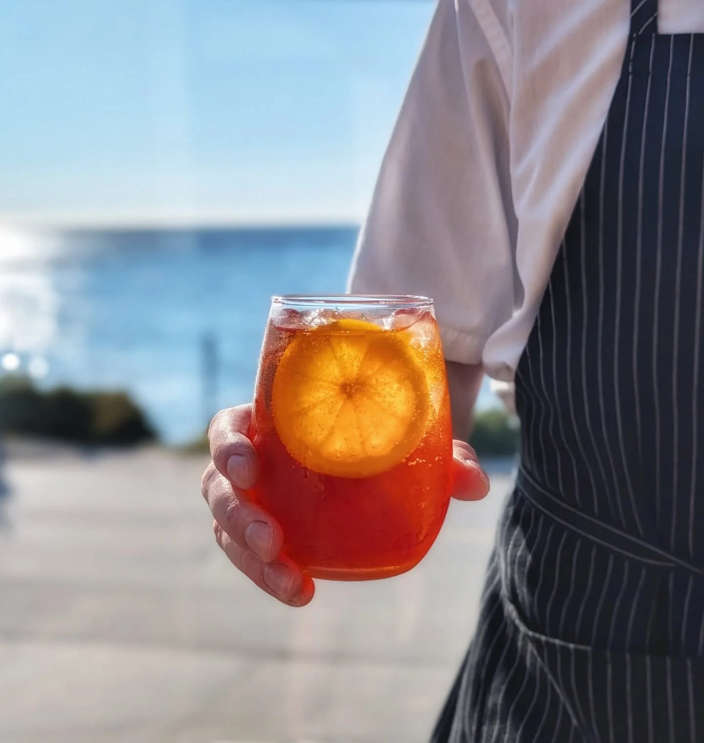 Person holding a glass of iced orange beverage with a lemon slice inside, near the water with a sunny sky.