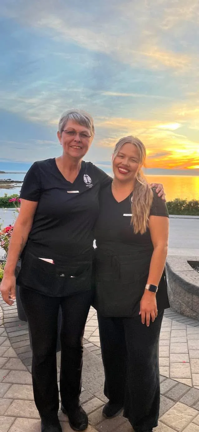 Two women wearing black uniforms standing arm-in-arm outdoors at sunset near a body of water, smiling.