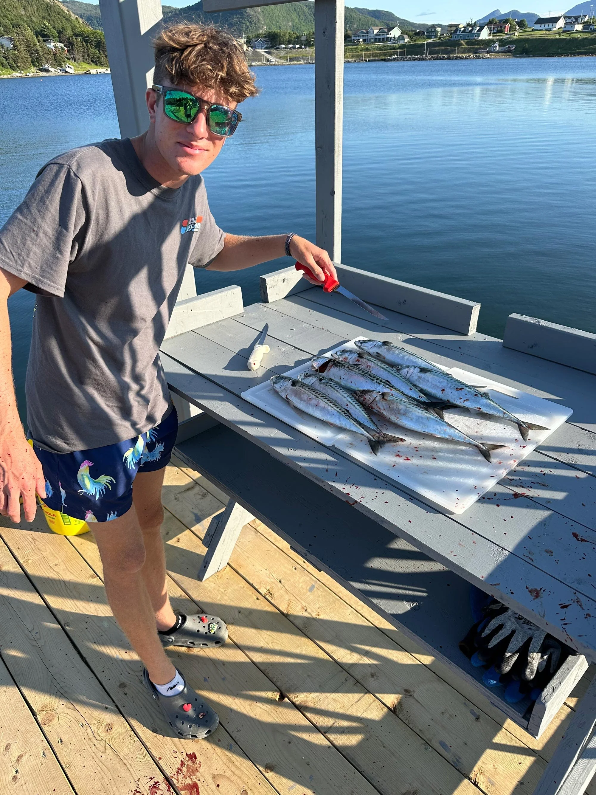 A young man with curly hair wearing sunglasses, a gray t-shirt, colorful shorts, and Crocs shoes, stands on a wooden dock next to a table holding several freshly caught fish. He is holding a red-handled knife and appears to be preparing or cleaning t