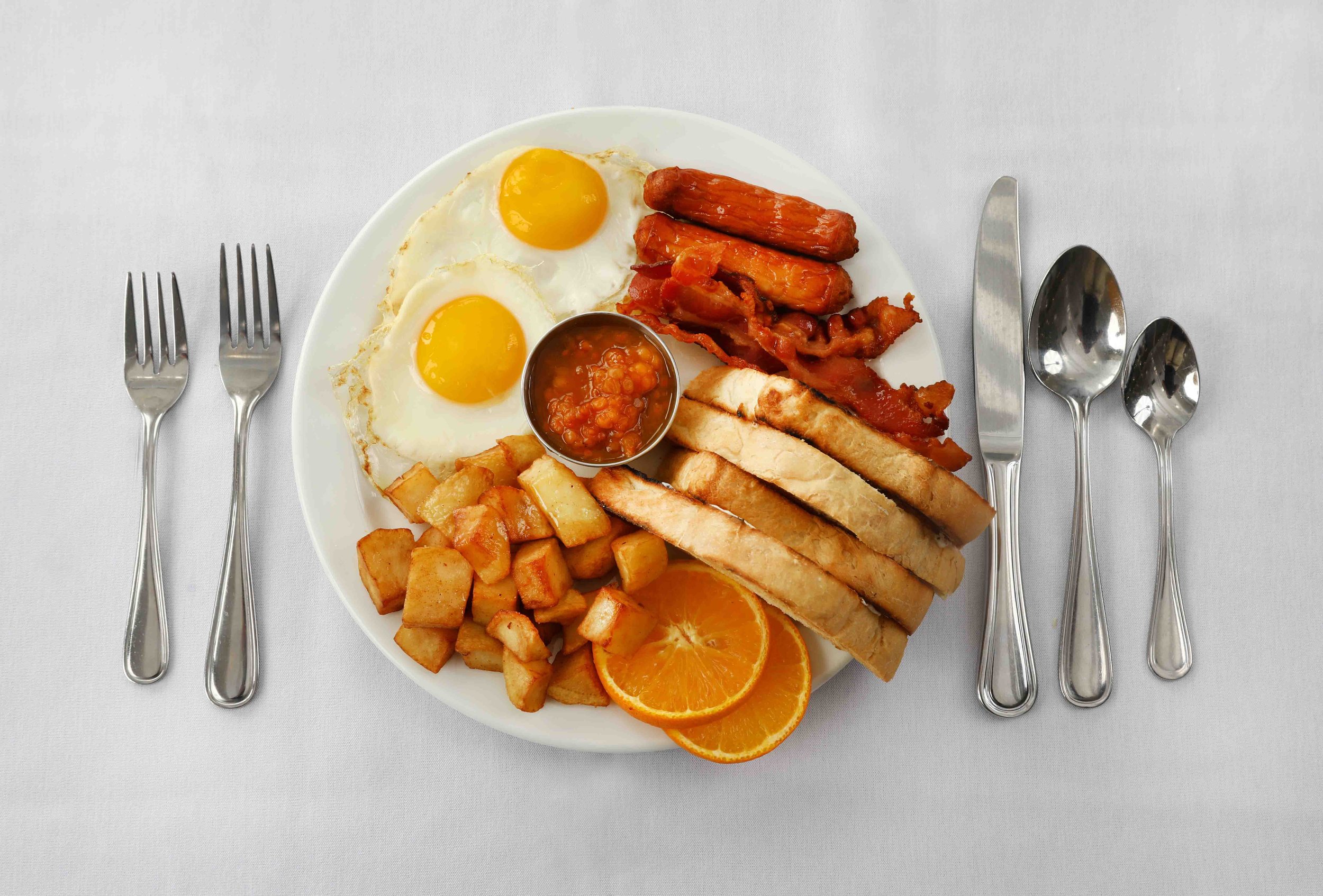 A plate of breakfast food with eggs, breakfast sausage, bacon, toast, breakfast potatoes, orange slices, and baked beans, set on a white tablecloth with utensils.