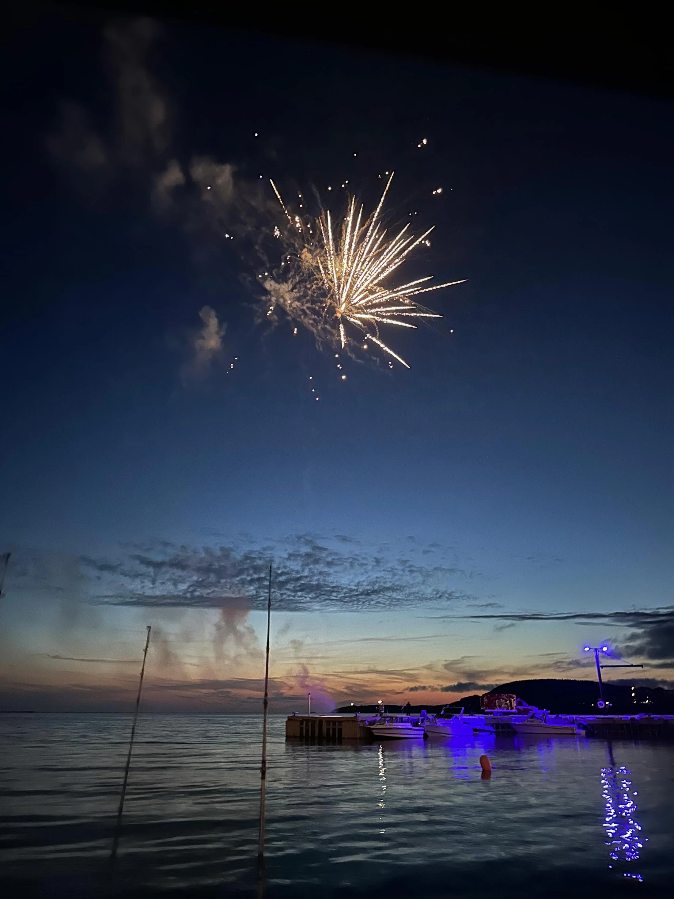 Fireworks display over a calm body of water at dusk, with boats docked near a pier and colorful lights reflecting on the water.
