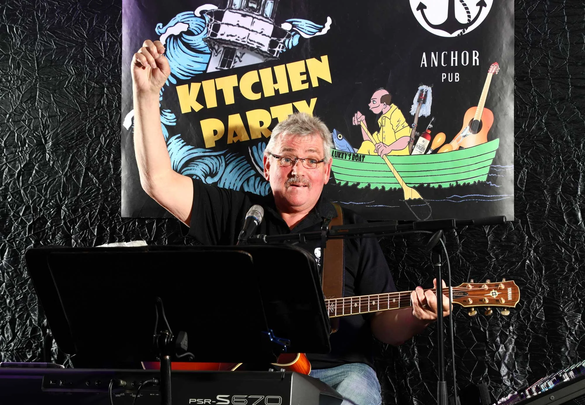 A man with glasses, a mustache, and gray hair is playing an electric keyboard and a guitar, raising his right arm with a fist in celebration or greeting, during a performance at a kitchen party event. There is a microphone in front of him. Behind him