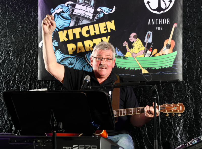 Man playing guitar and singing at a kitchen party event with a sign in the background reading "Kitchen Party" at Anchor Pub.