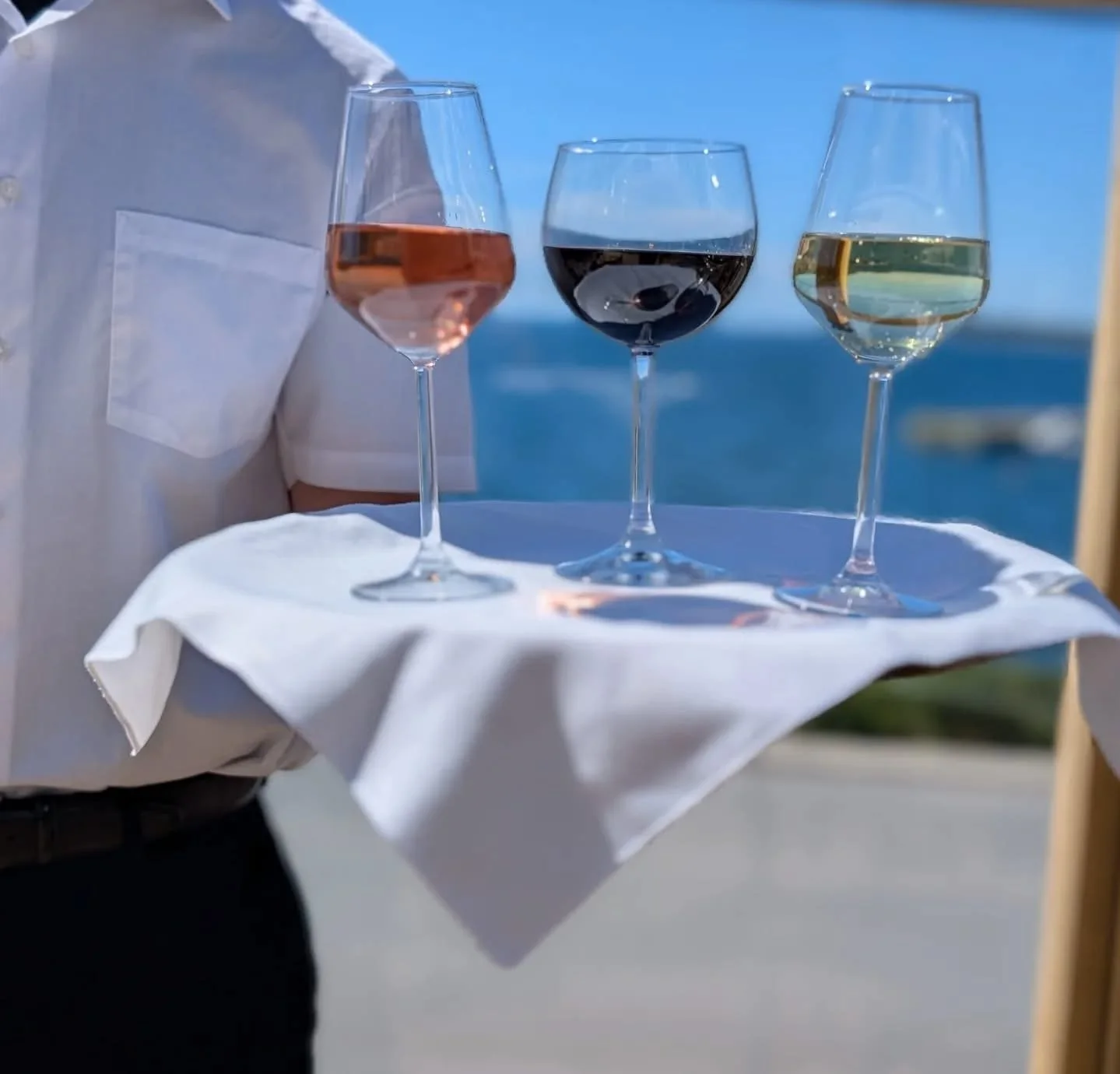 A server holding a white cloth with three wine glasses filled with different colored wines, against a background of a blue sky and ocean.