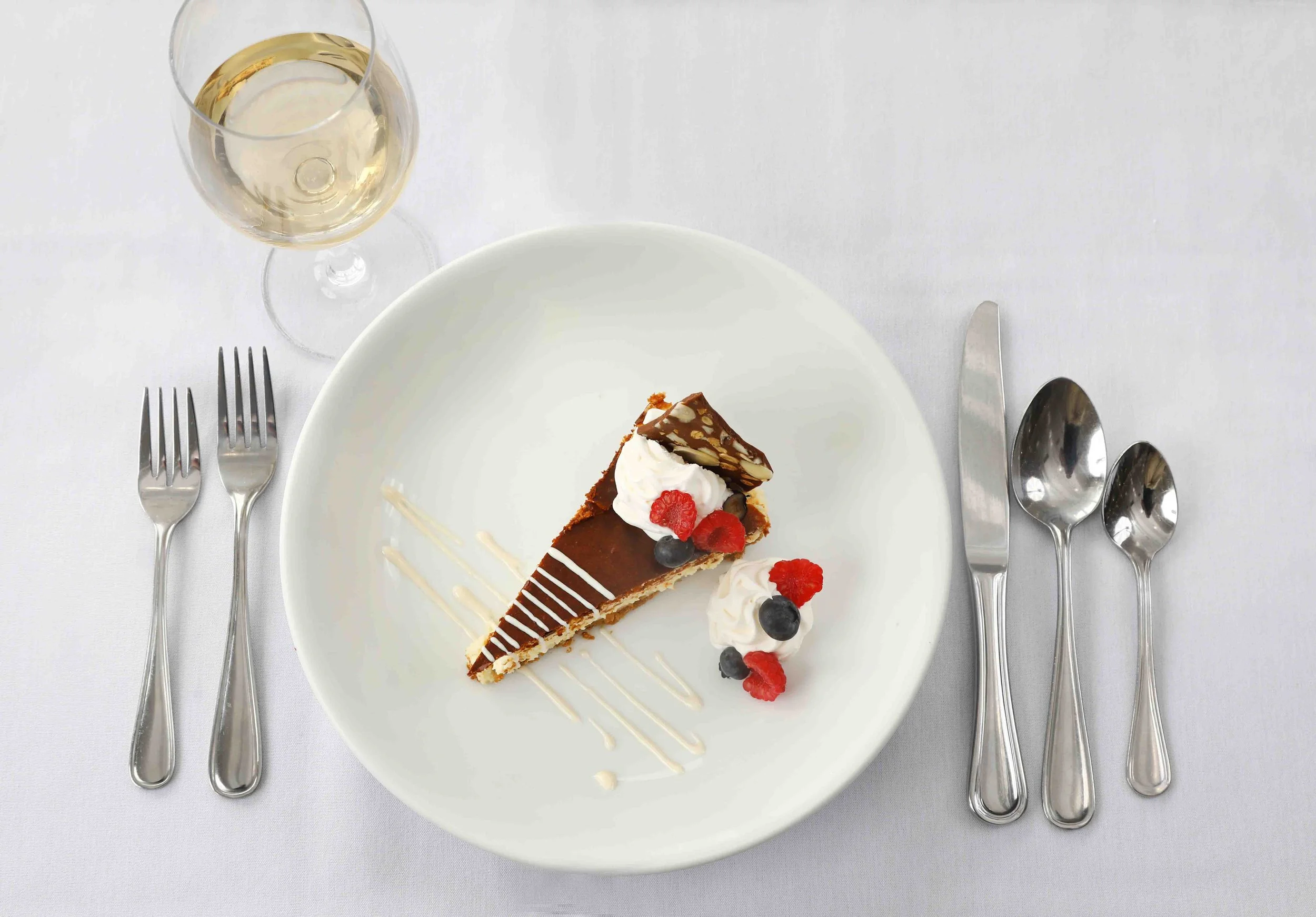A dessert plate with a slice of chocolate cake garnished with whipped cream, fresh raspberries, and blueberries, on a white tablecloth, with a glass of white wine to the top left and silver cutlery on each side.