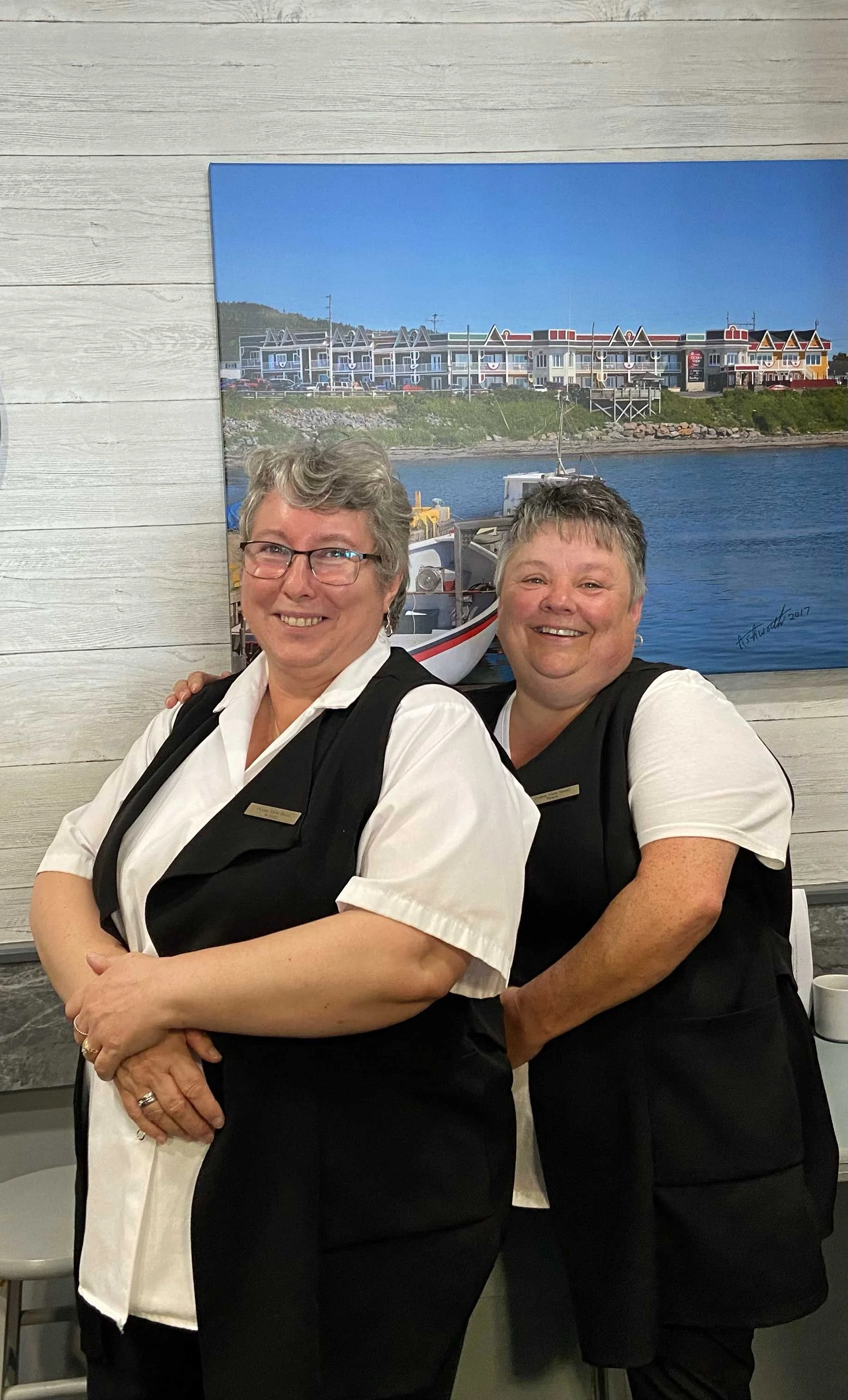 Two smiling women wearing black aprons, standing in front of a wall with a large painting of a harbor scene.