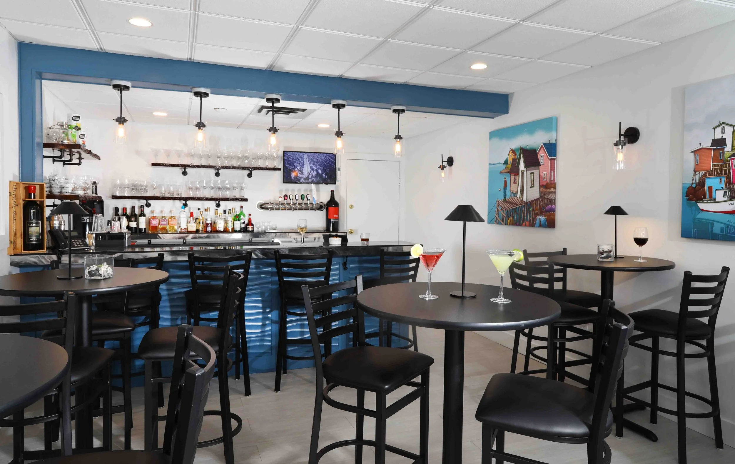 Interior of a modern bar and lounge area with black tables and chairs, colorful drinks on the tables, paintings on the wall, and a bar counter with hanging glasses and alcohol bottles.