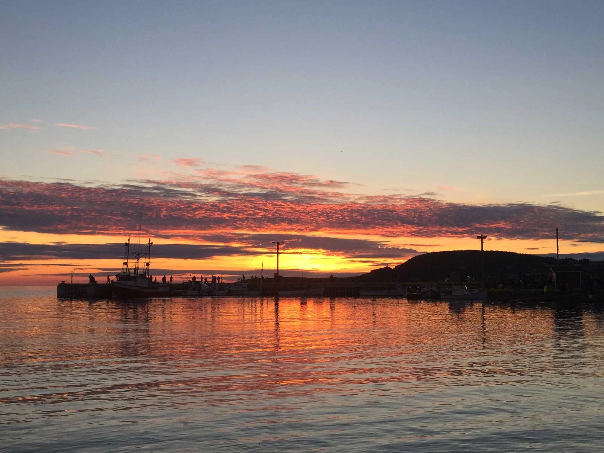 Sunset over a harbor with boats docked along a pier, pink and orange clouds in the sky, and the calm water reflecting the colors.