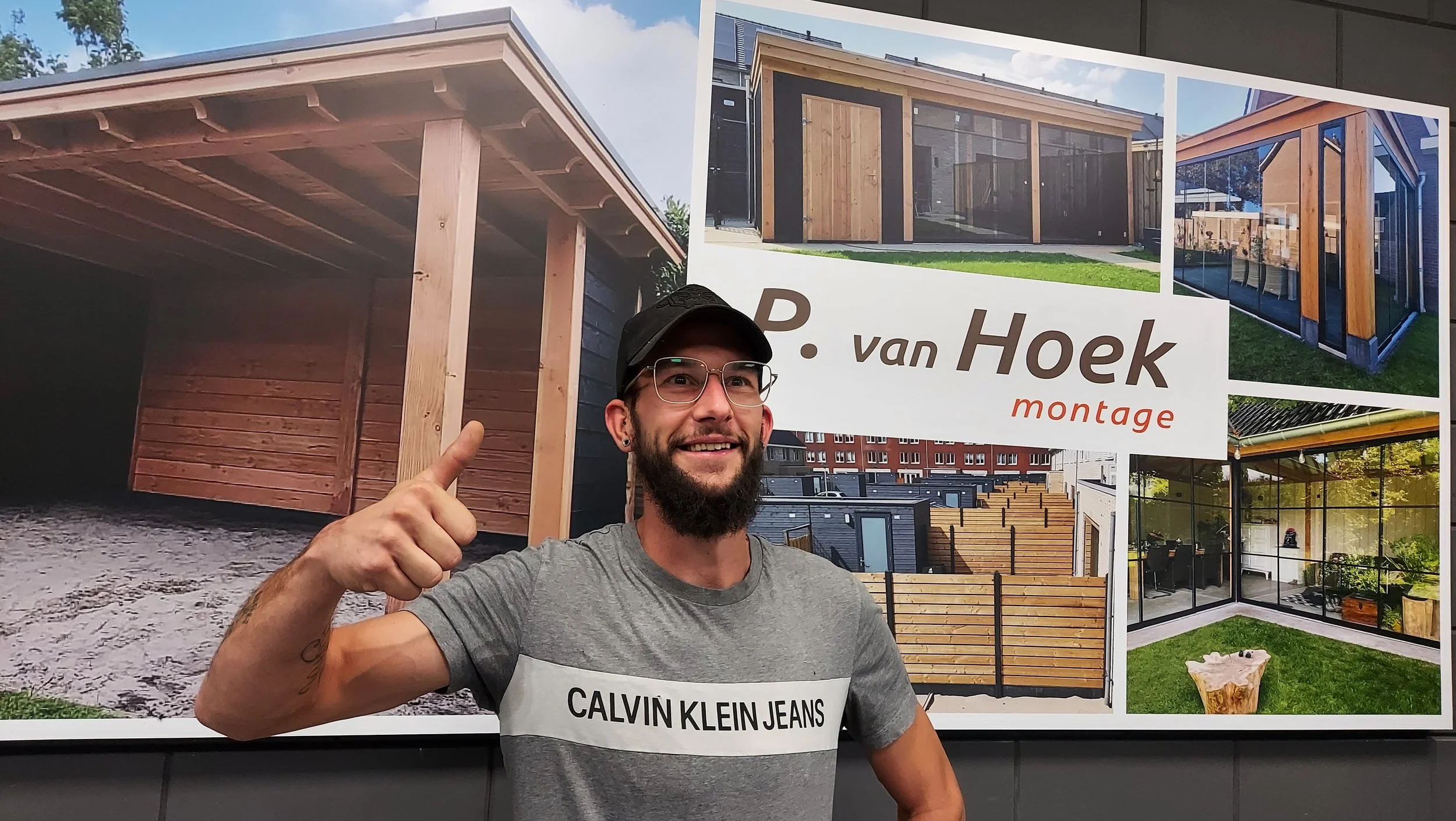 Man met bril, pet en grijze Calvin Klein T-shirt geven duim omhoog in voorgrond, achter hem een grote bouwstijl poster met verschillende huizen en bouwprojecten.