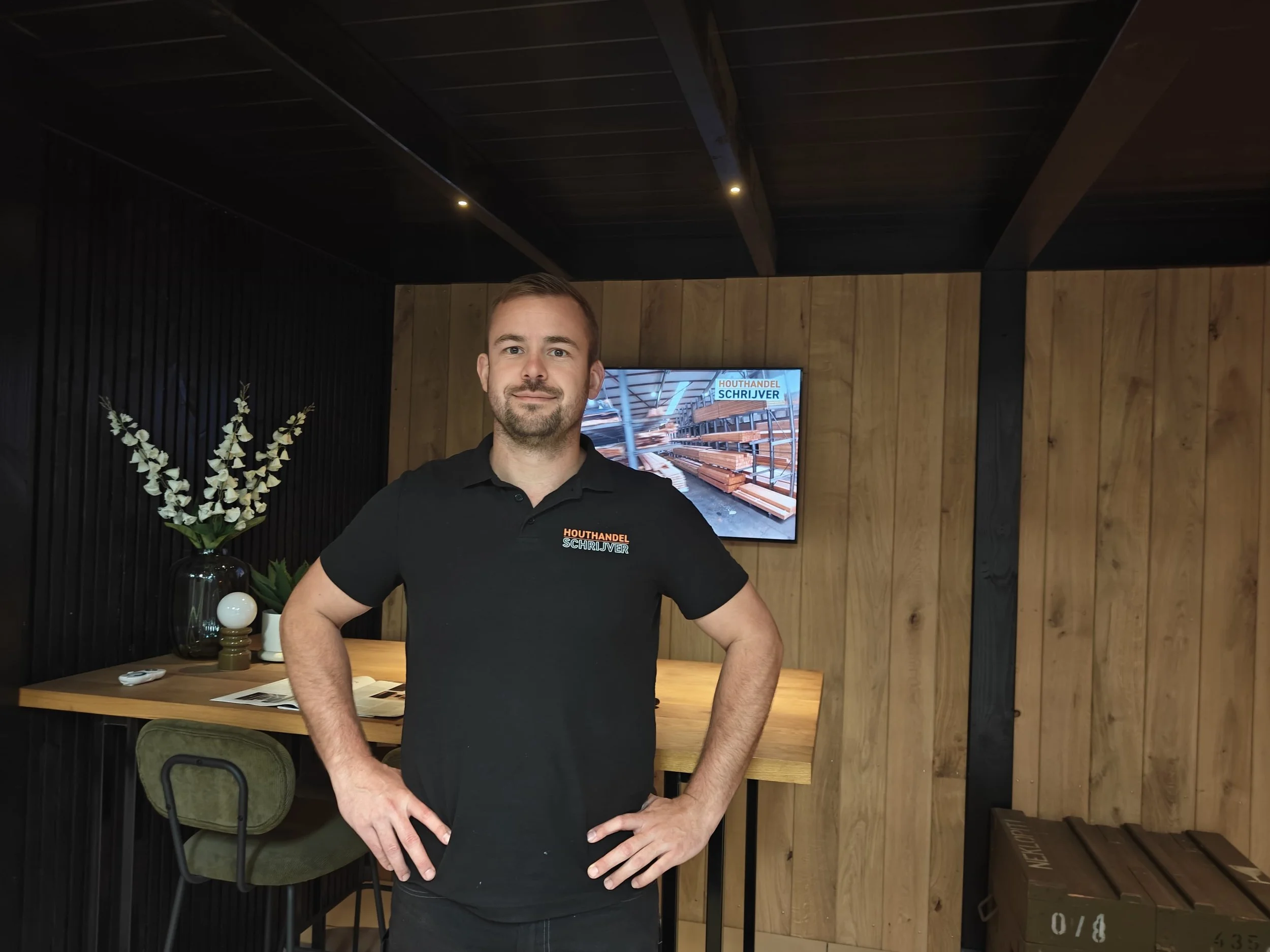 Man staat binnen bij Houthandel Schrijver, draagt een zwart polo shirt met het logo van het bedrijf, naast een tafel met een boek en decoratieve objecten en er is een tv aan de muur die een afbeelding van een houtwinkel toont.