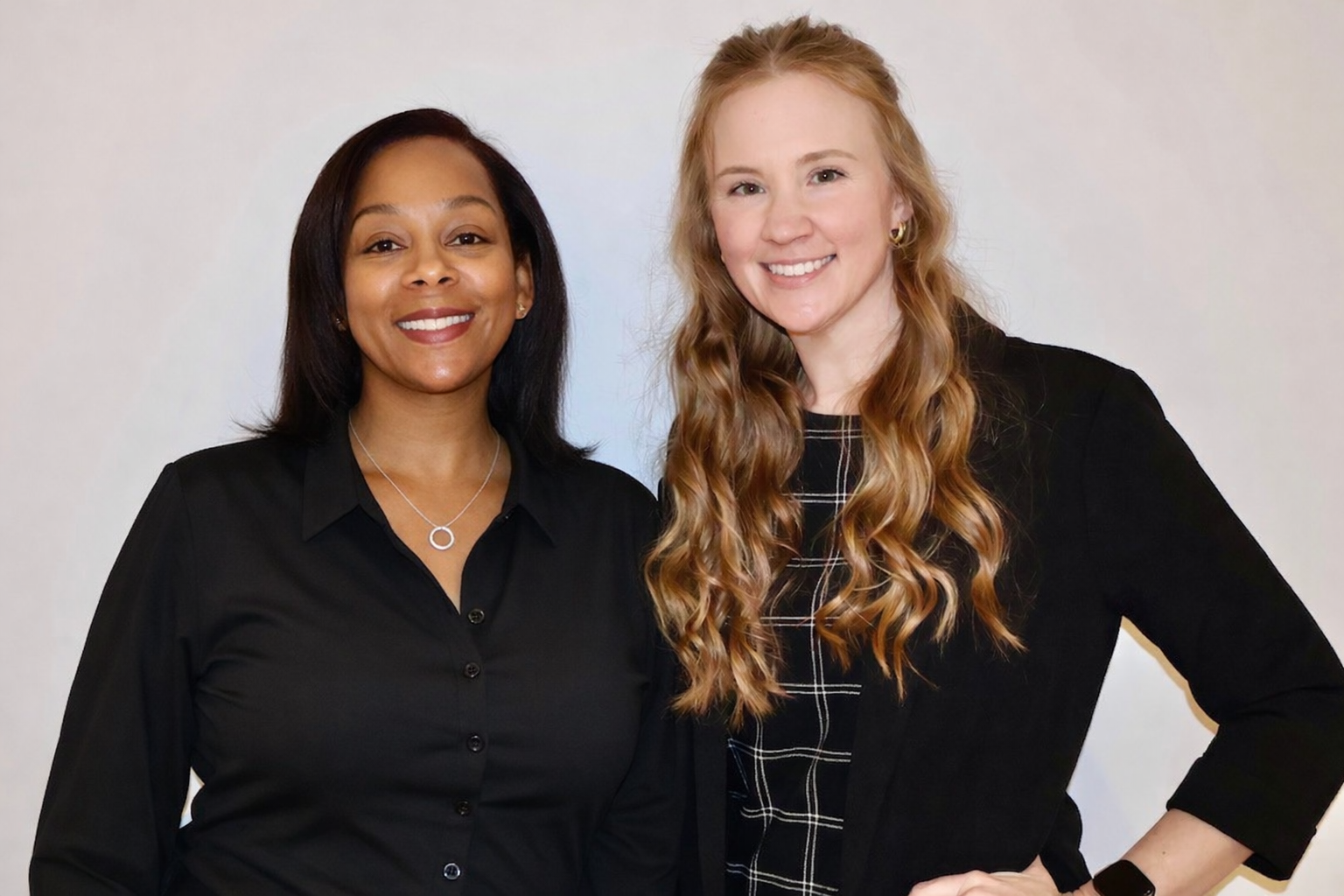 Two smiling women standing side by side, wearing black clothing against a light-colored background.