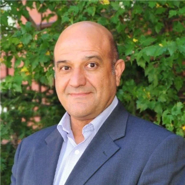 A man in a dark suit and white shirt standing outdoors with green leafy trees in the background.