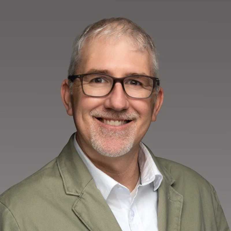 A smiling middle-aged man with glasses, gray hair, and a gray beard, wearing a beige blazer and a white shirt, against a gray background.