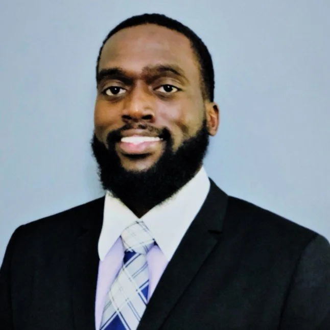 Portrait of a smiling Black man in a business suit with a white shirt and a purple plaid tie, standing against a plain blue background.