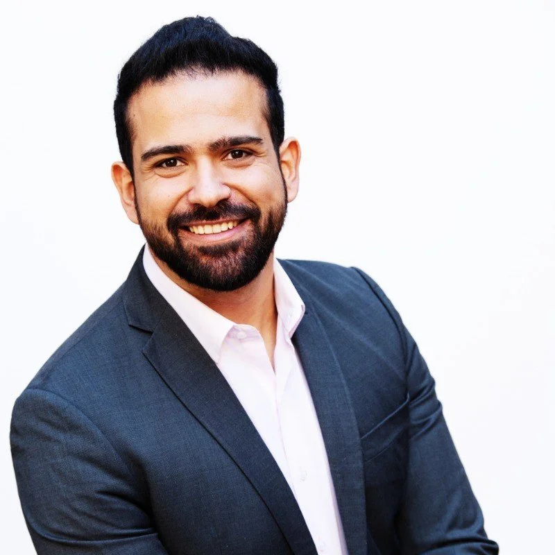Professional portrait of a man with dark hair and beard, smiling, wearing a navy blazer and white shirt, against a plain white background.