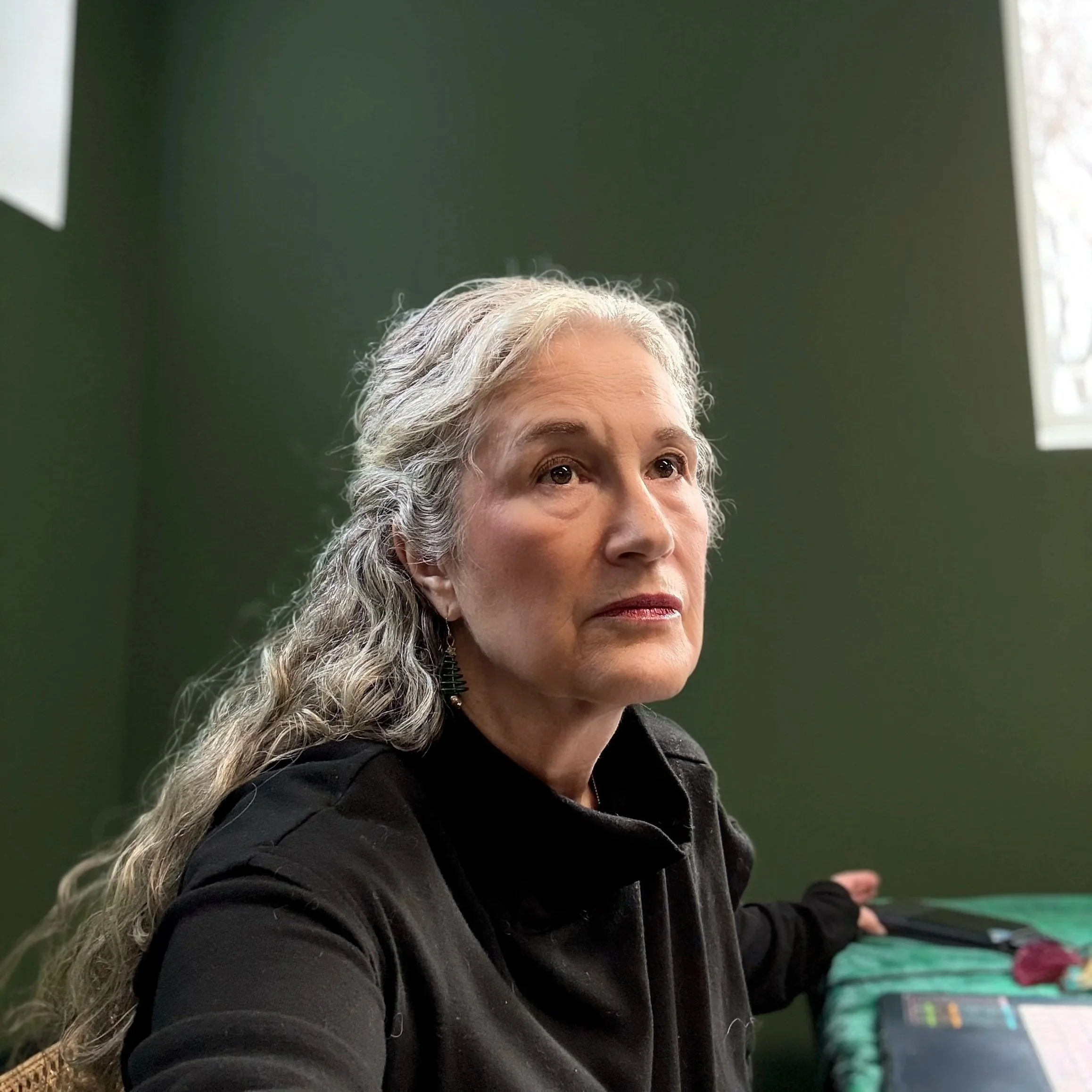 Close-up of an older woman with gray curly hair, wearing a black jacket, sitting indoors in front of a green wall corner, with 1 window on each wall.