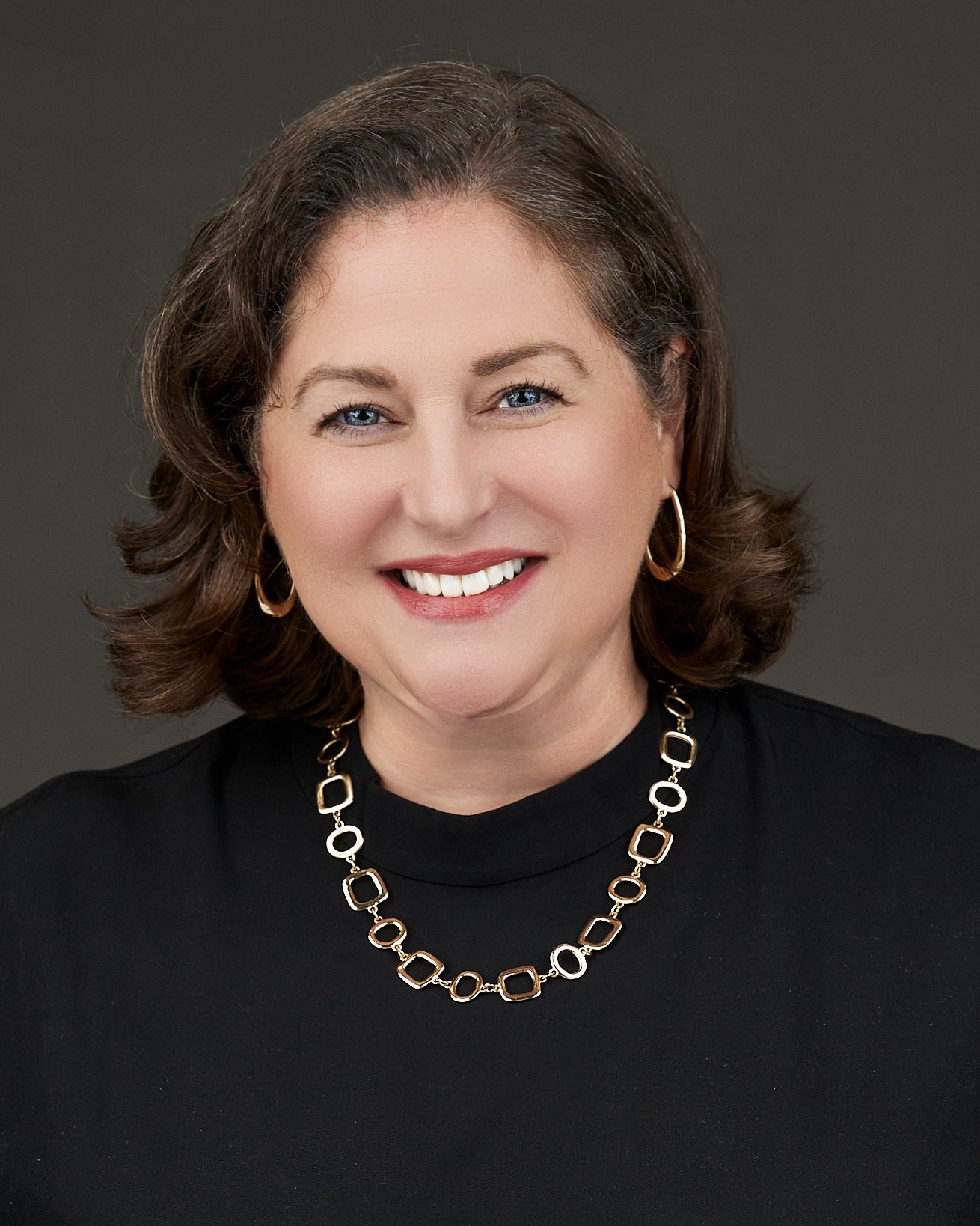 A woman with shoulder-length brown hair, blue eyes, and wearing a black top, gold hoop earrings, and a gold and black geometric necklace, smiling at the camera against a gray background.