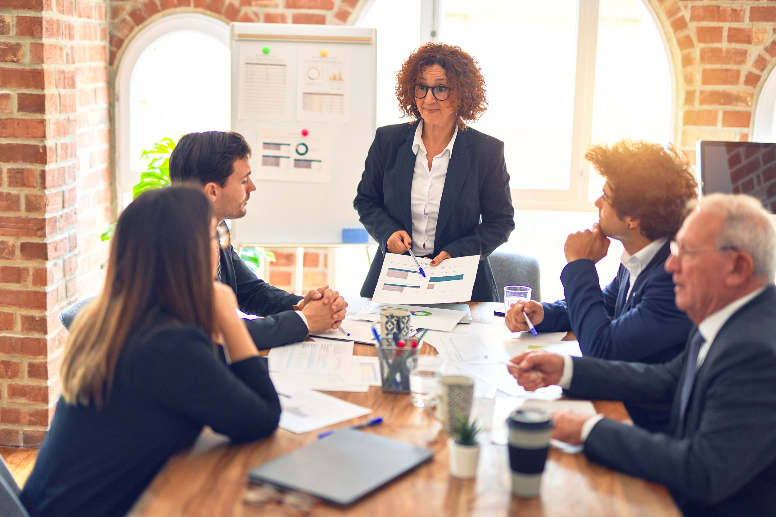 A woman in a black blazer and glasses is standing in front of a group of business professionals seated at a conference table, presenting reports and charts in a well-lit room with brick walls and large windows.