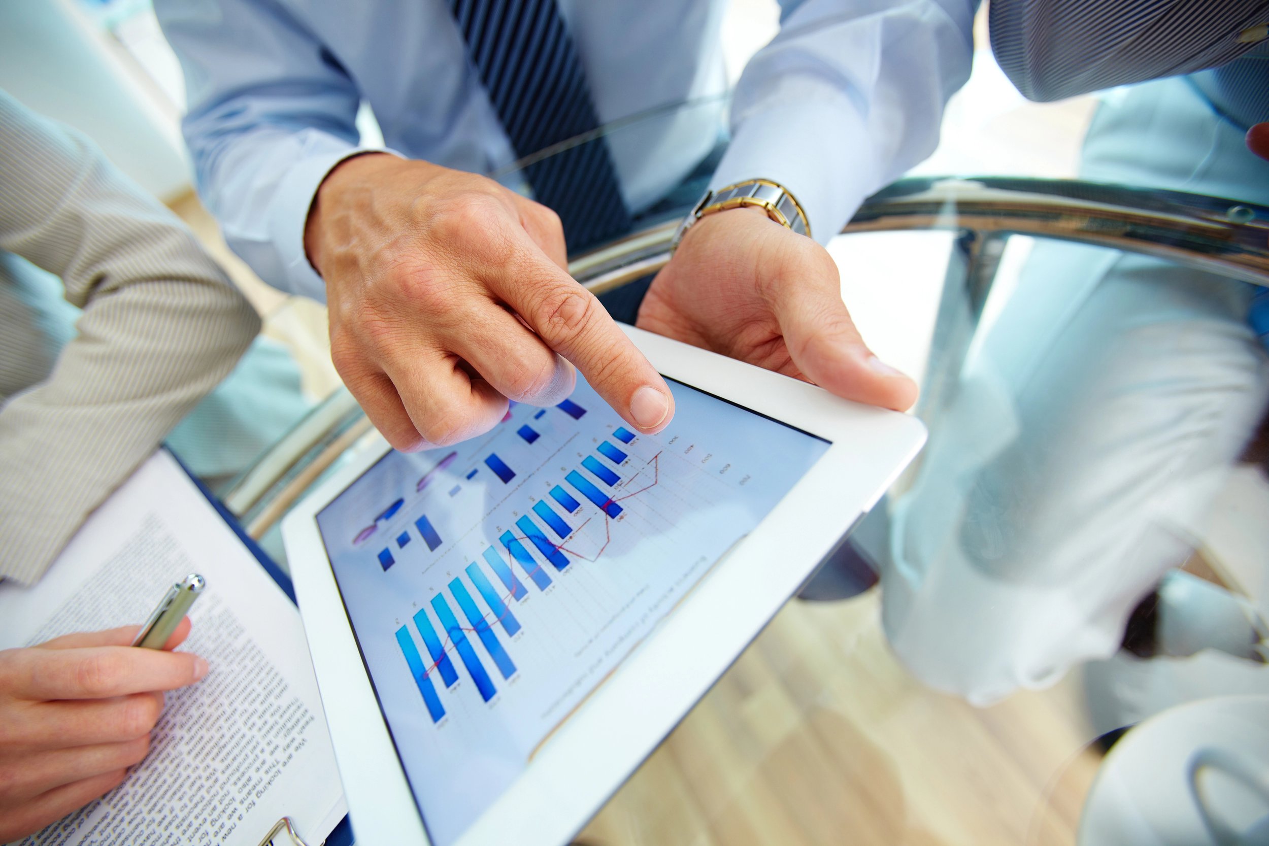 Multiple people in business attire analyzing data on a tablet device with charts and graphs, possibly in a meeting or presentation setting.