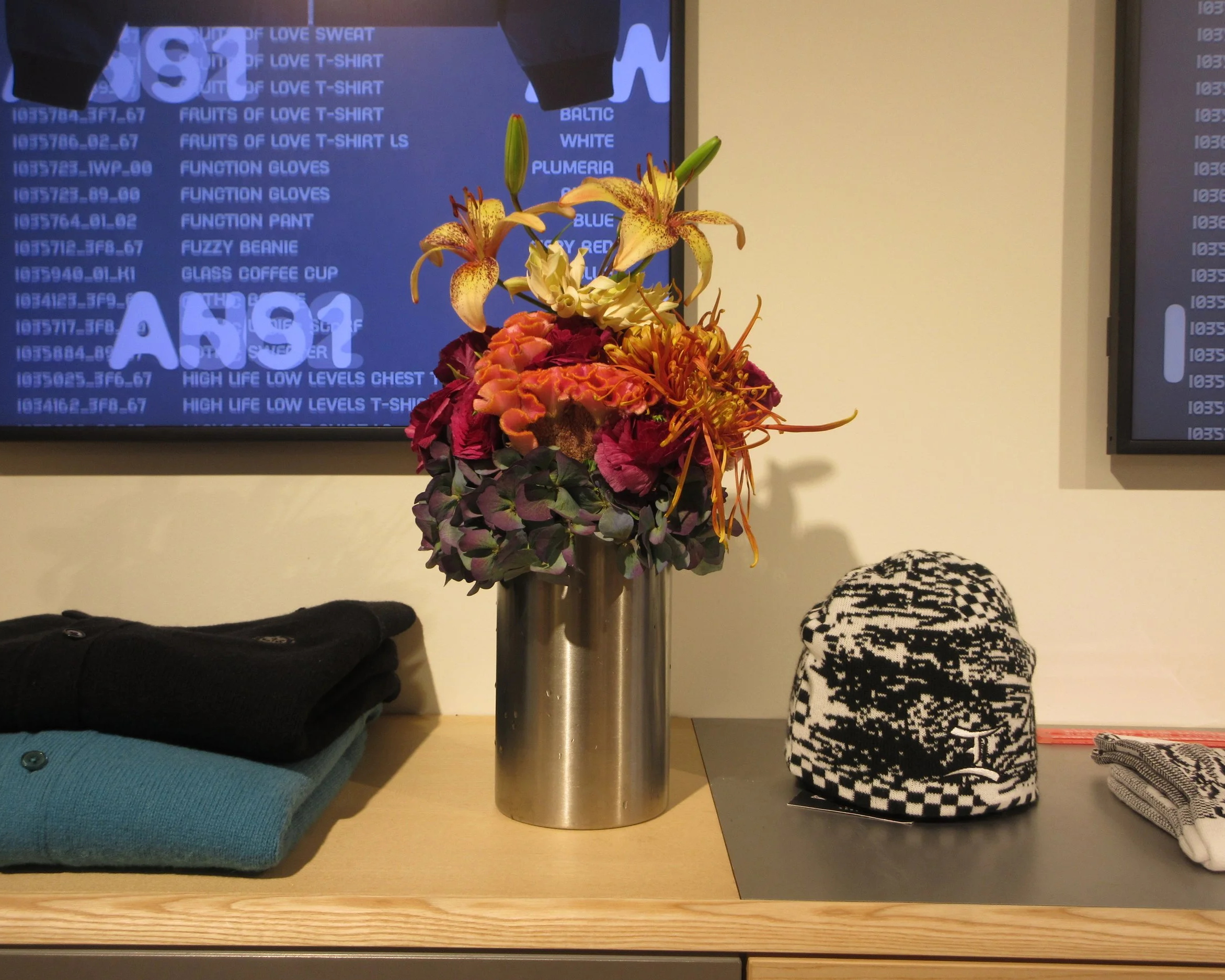 Vase avec fleurs colorées, téléviseurs à l'arrière, vêtements empilés et casquette à motifs noir et blanc sur une table.