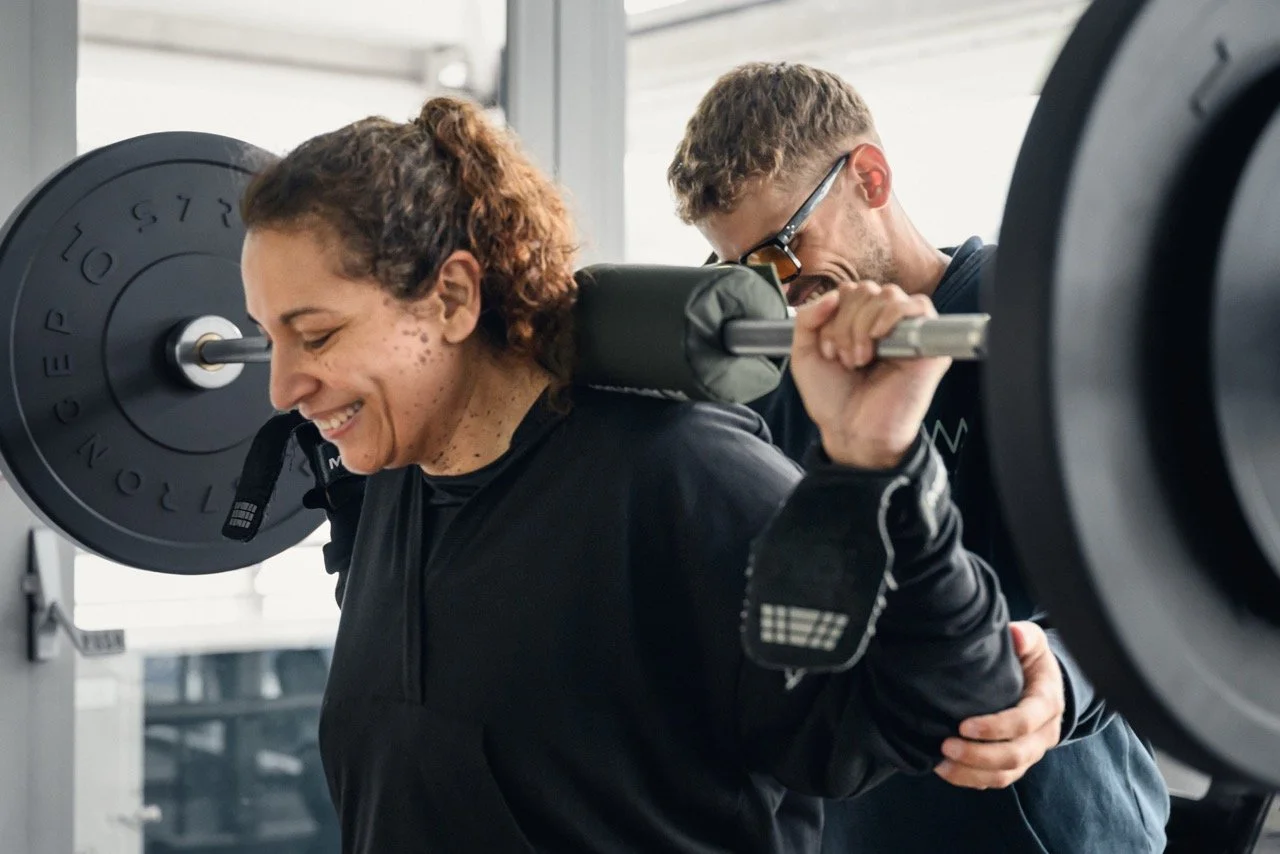 Woman smiling while doing a squat with a barbell on her shoulders, assisted by personal trainer